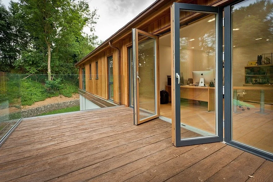 Open glass doors leading from a wooden deck to a modern office with a computer and shelves inside.