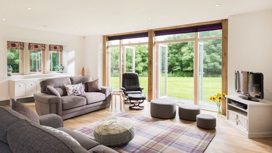 Bright living room with large windows and a view of green outdoor space, furnished with sofas, an office chair, small ottomans, a TV on a white stand, and a sunflowers in a vase.