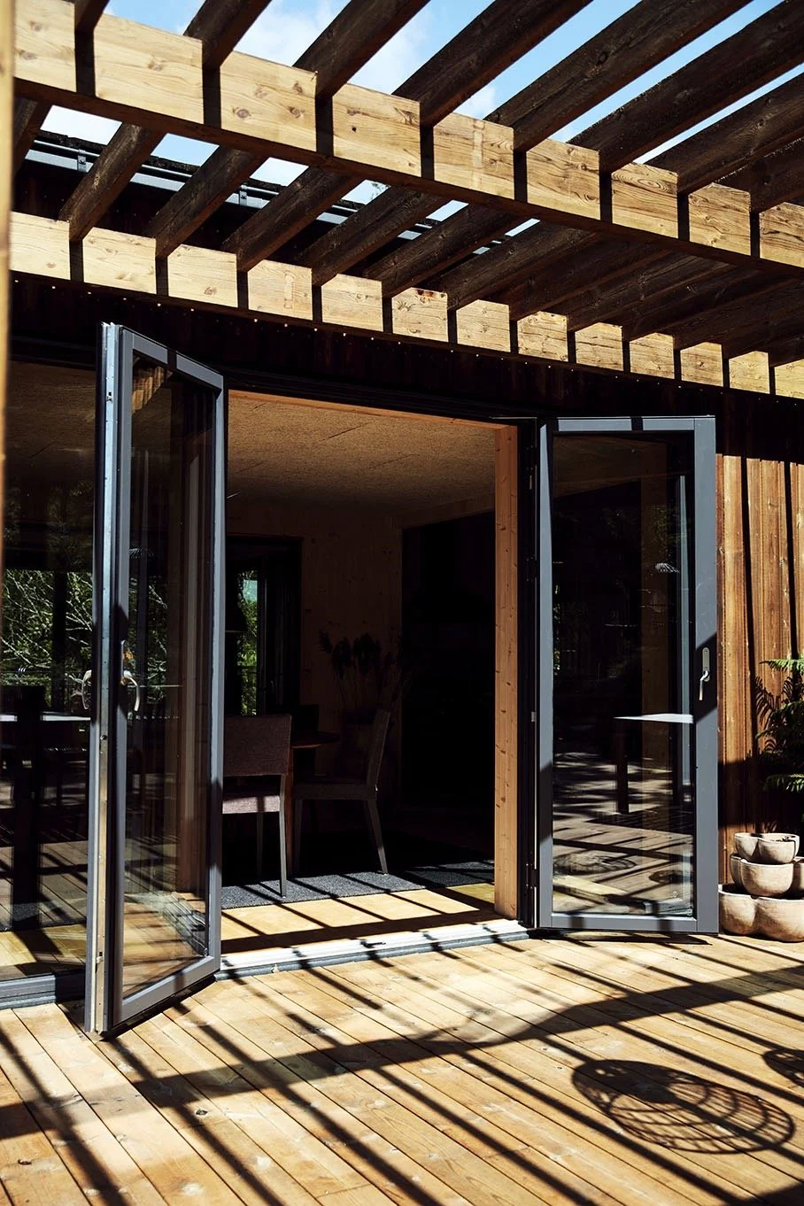 A wooden deck with open glass sliding doors leading into a room with chairs and a table. Wooden beams and a wooden railing are above the deck, with shadows cast on the floor. Potted plants are visible on the right side.