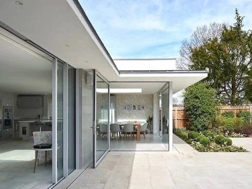 View of an modern house with large glass sliding doors, an indoor dining area, and a garden outside with trees and shrubs.