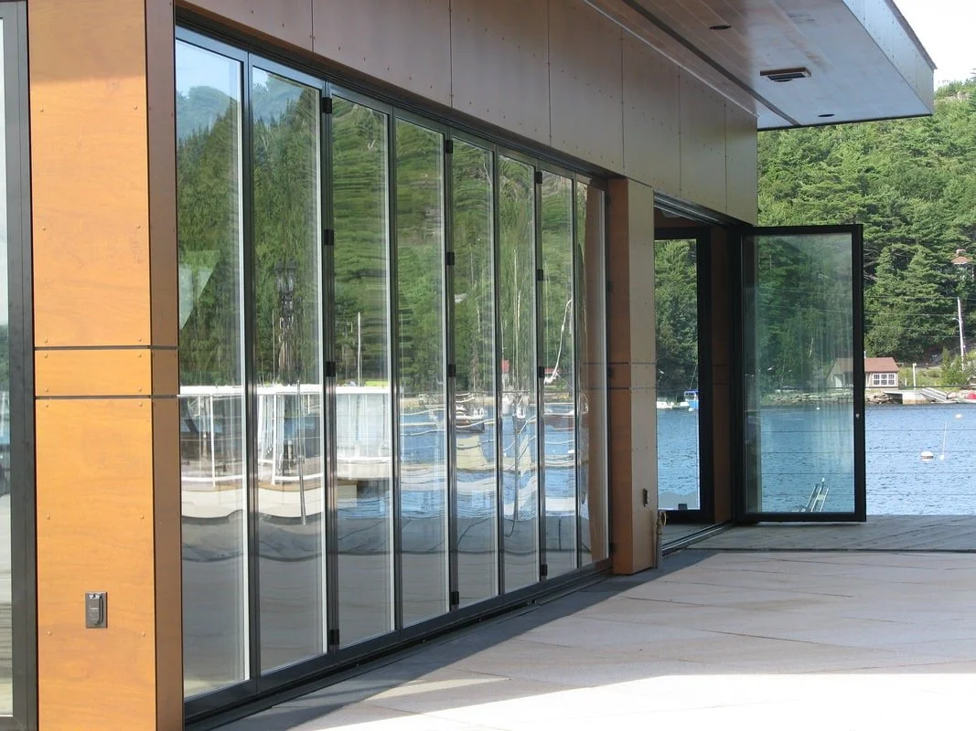 Modern house with glass sliding doors overlooking a lake and boats, greenery in the background.