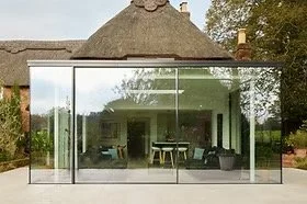 Modern glass house with large glass walls showing interior living space and outdoor greenery