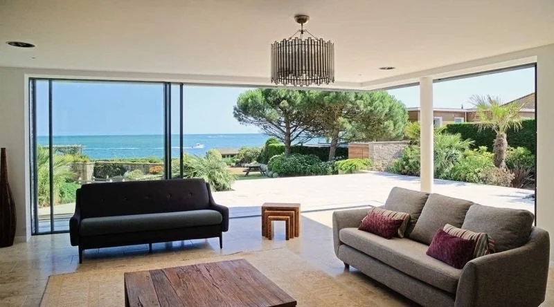 Living room with large glass windows showing ocean view, with two sofas, a wooden coffee table, and a chandelier.