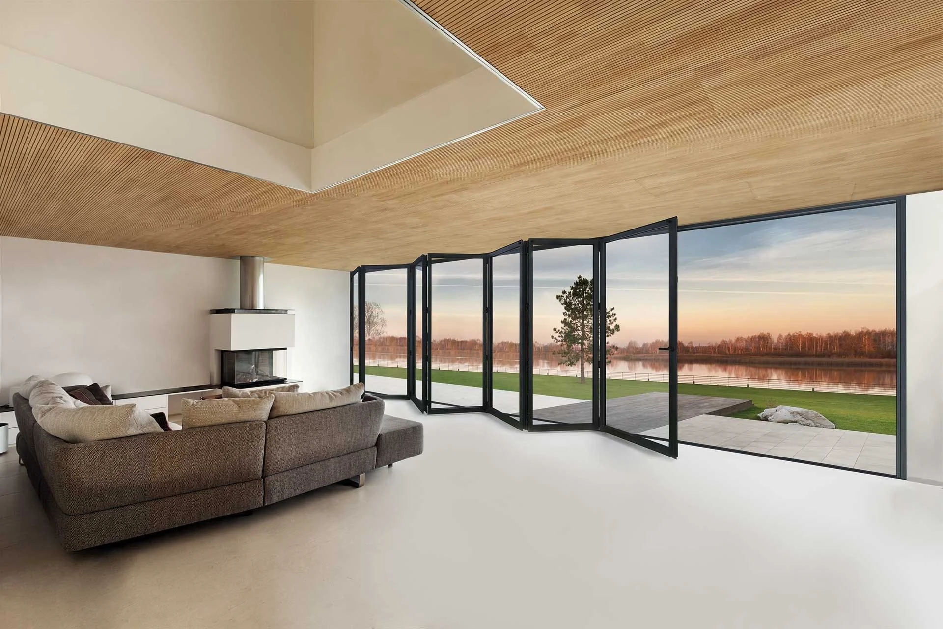 Modern living room with large black-framed folding glass doors opening to an outdoor patio and scenic lake view at sunset, featuring wooden ceiling accents and a cozy sectional sofa.