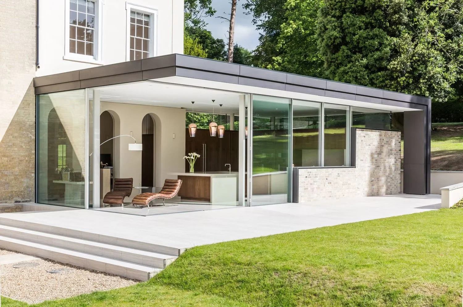 Modern glass house extension with outdoor seating area, green lawn, and trees in background