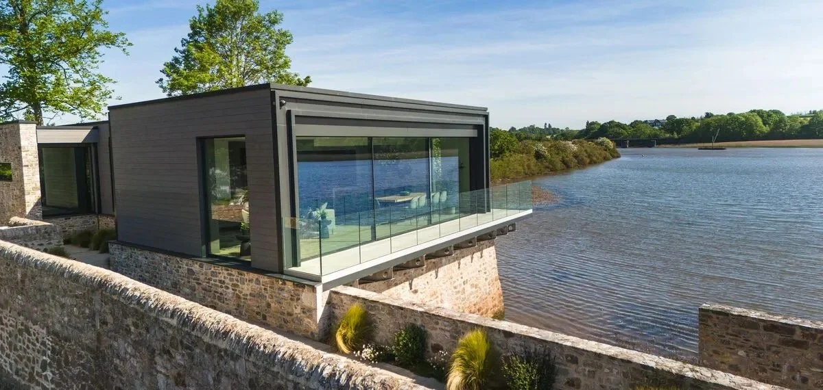 Modern house with large glass windows and a glass balcony, built on a stone foundation, overlooking a body of water with green trees in the background.