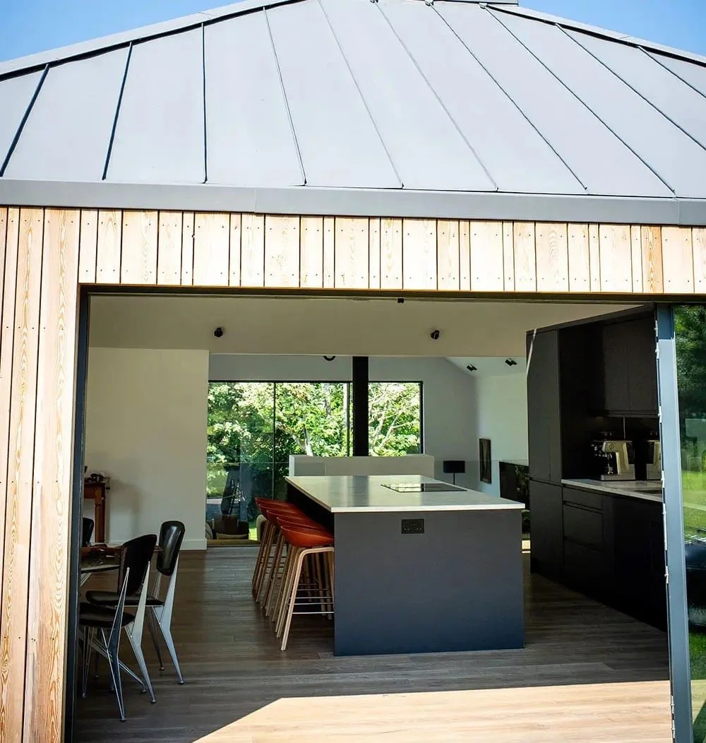 Modern kitchen with black cabinets, an island with barstools, and large windows showing greenery outside.
