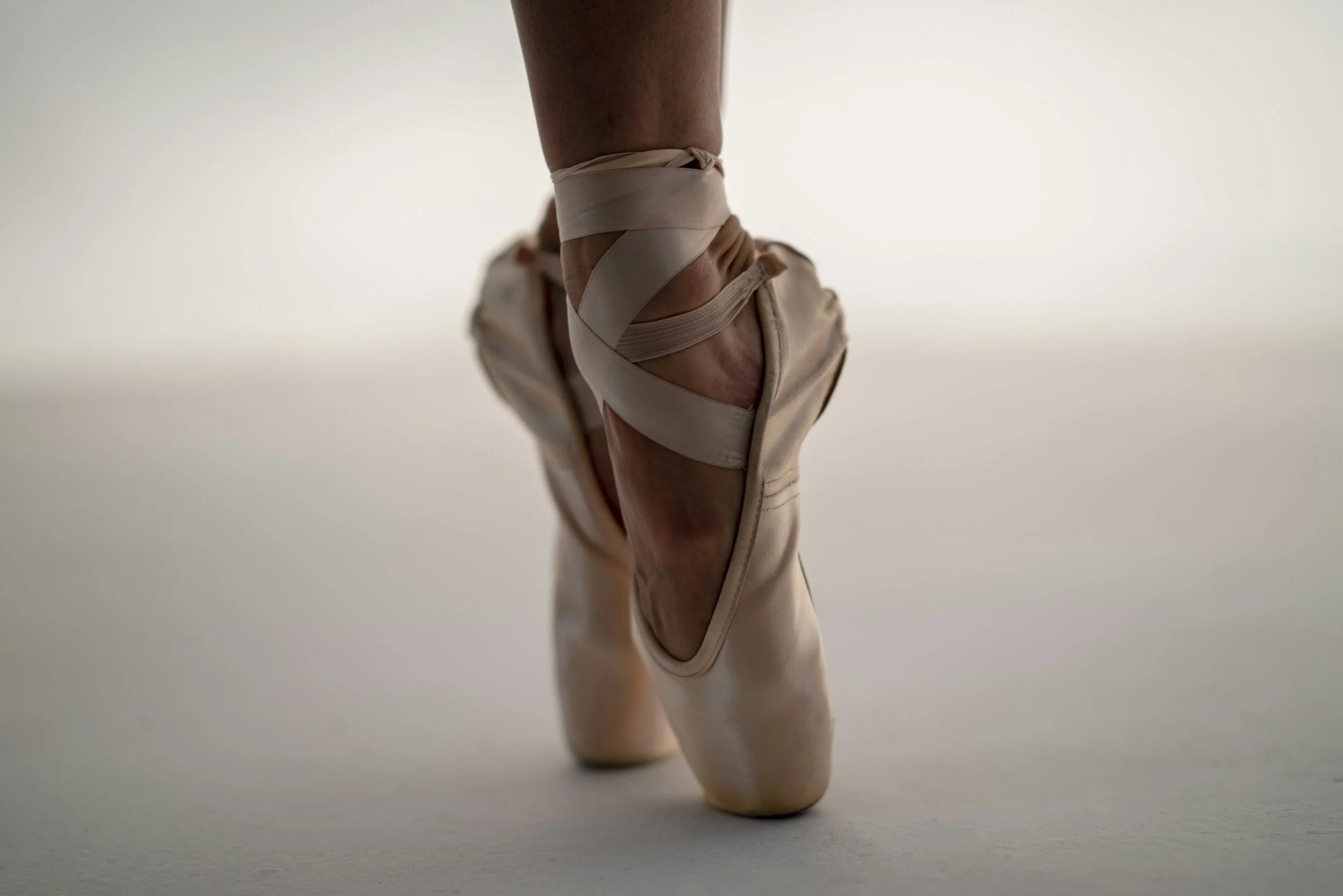 Close-up of ballet pointe shoes worn by a dancer, with the dancer standing on their toes on a plain surface.