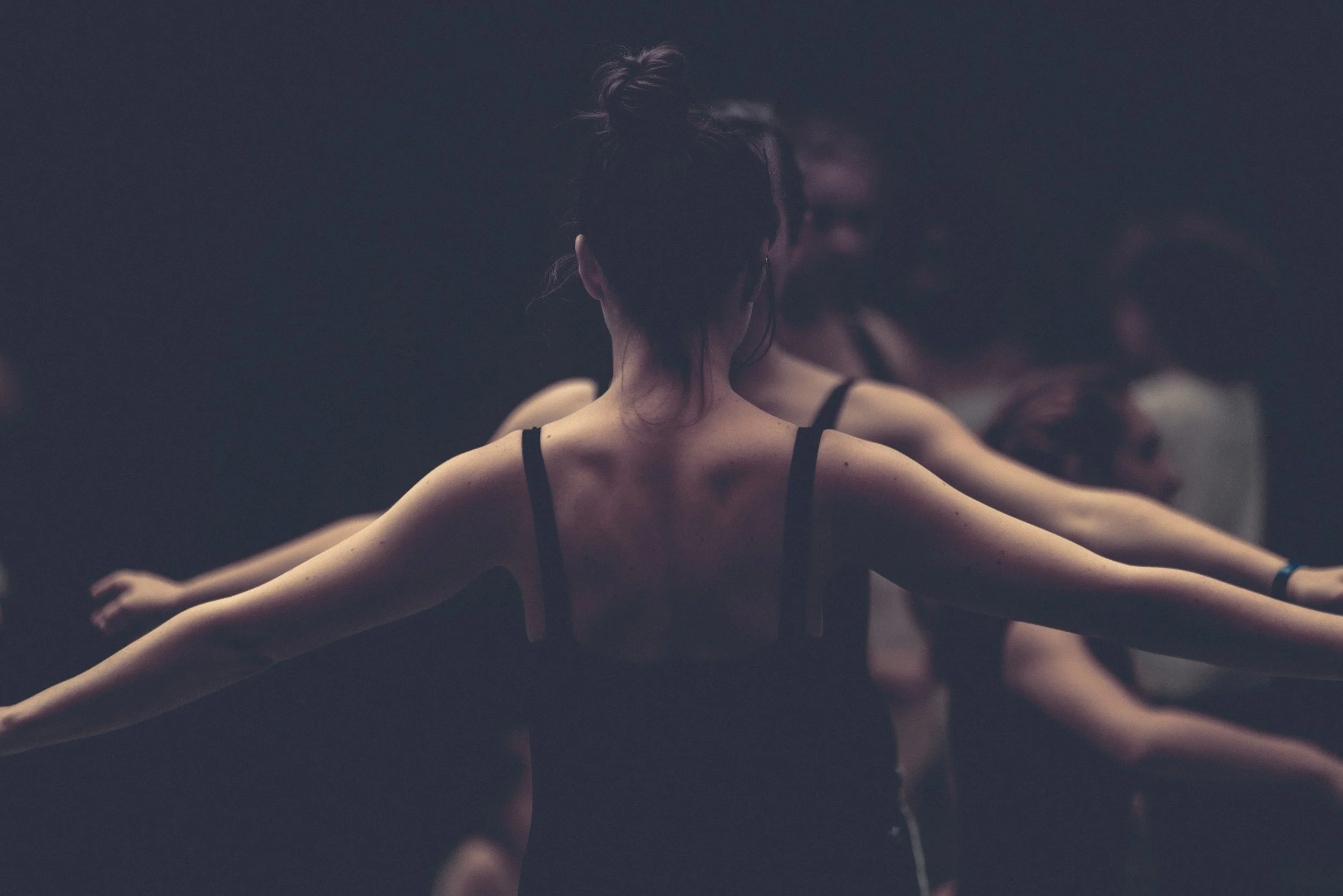 A person with a bun hairstyle wearing a black tank top practicing ballet with arms outstretched. Mirror reflection visible in a dark dance studio.