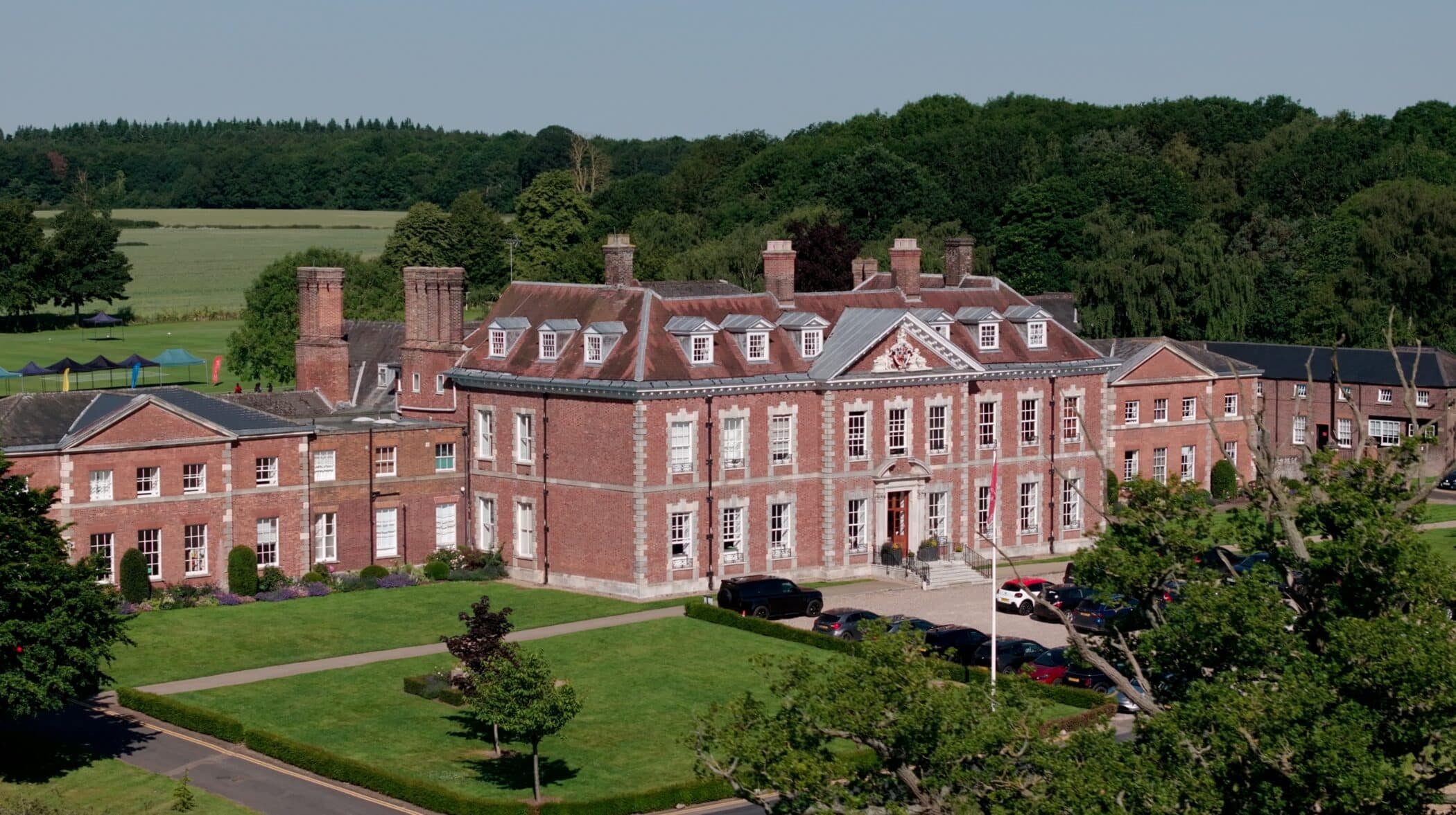 A large brick building with classical architecture, multiple chimneys, a manicured lawn, and a parking lot in front. Green trees and hills are in the background.