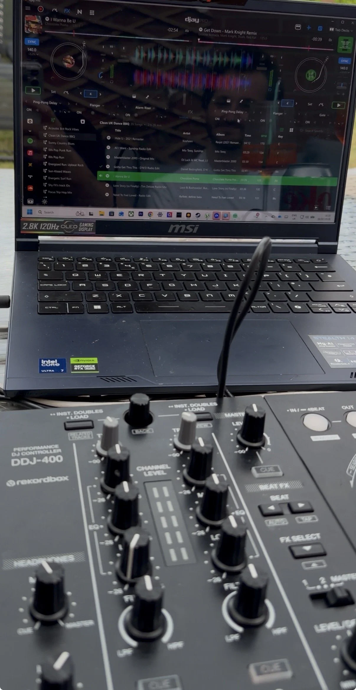 Close-up of a DJ mixing console with various knobs and sliders, with a laptop displaying a DJ software interface in the background.