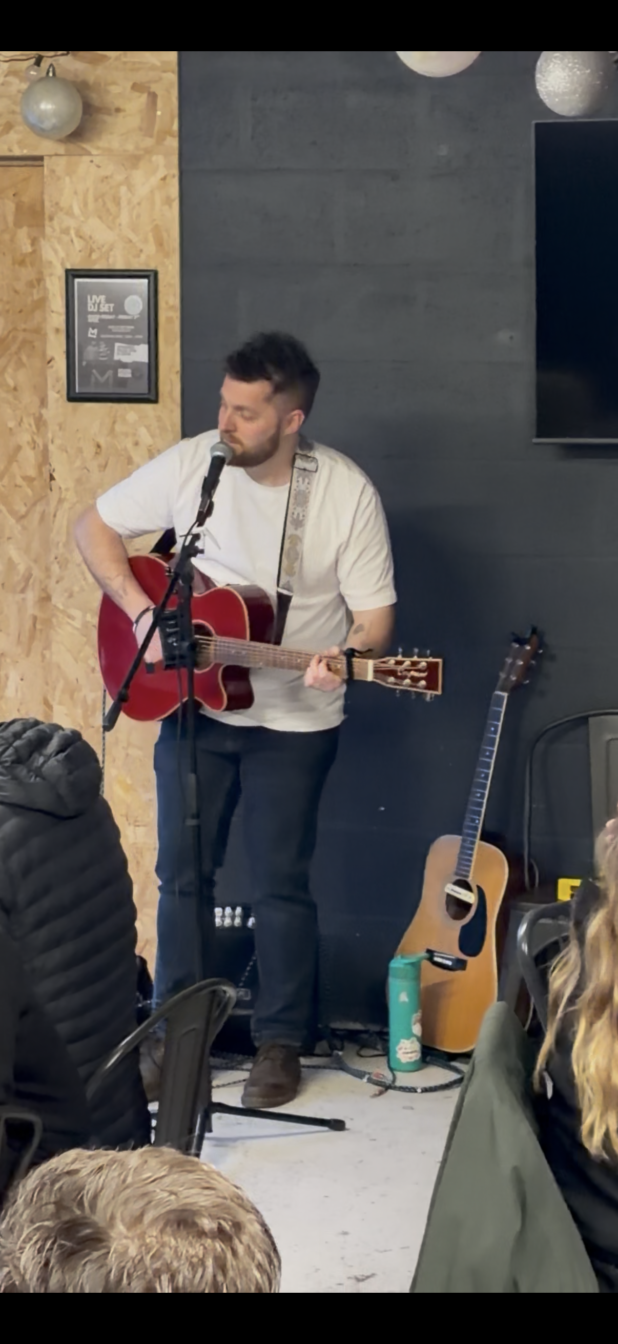 A man with dark hair and a beard is playing a red acoustic guitar and singing into a microphone. He is wearing a white shirt and standing in an indoor space with a gray wall and wood paneling. There is an additional guitar resting on a stand nearby and some personal items on the ground.