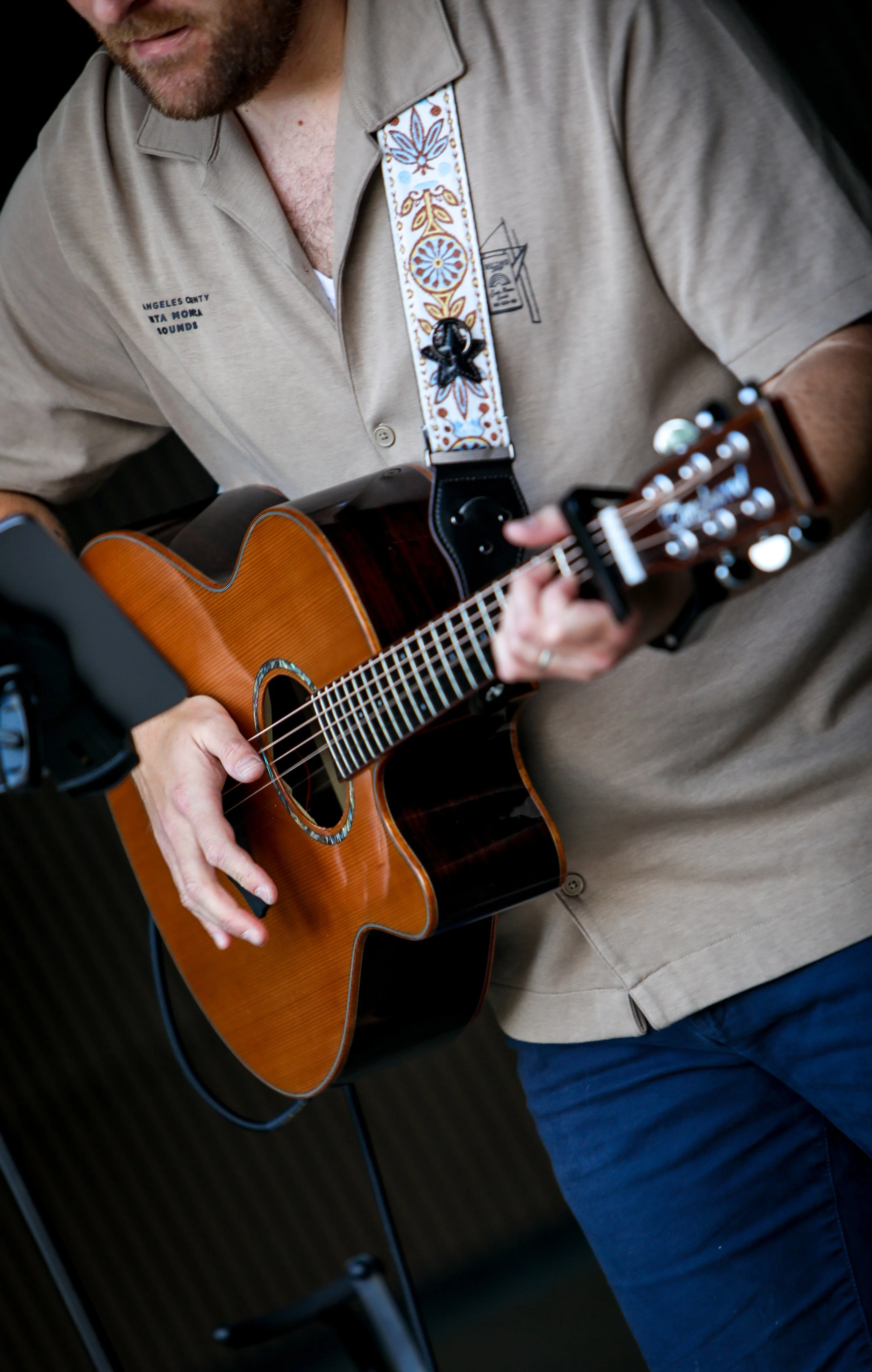 A man playing an acoustic guitar, wearing a beige shirt with a decorative strap and a watch, with a dark background.