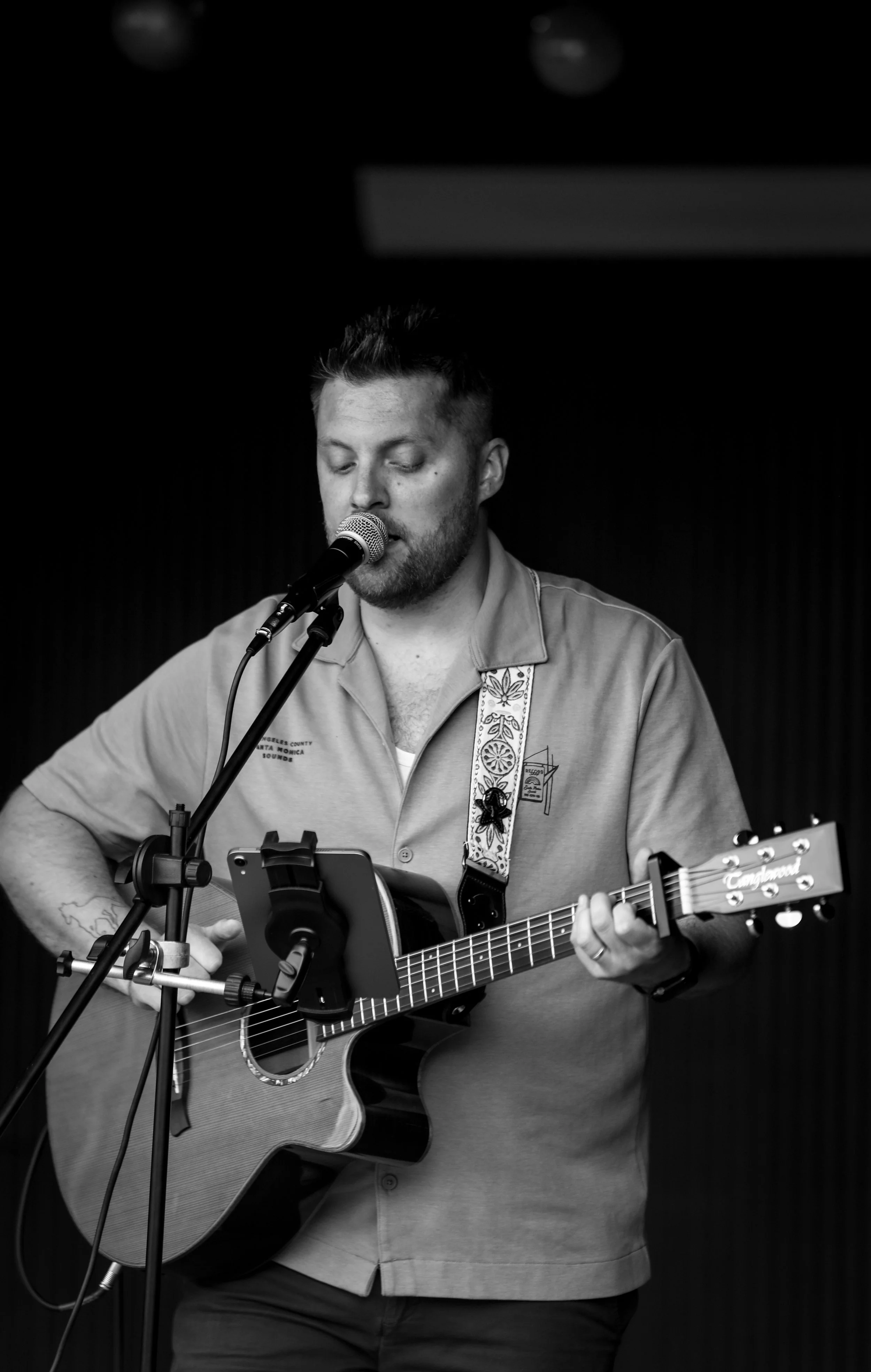 A man with short dark hair and stubble, wearing a light-colored shirt with text, playing an acoustic guitar and singing into a microphone in a dark room.