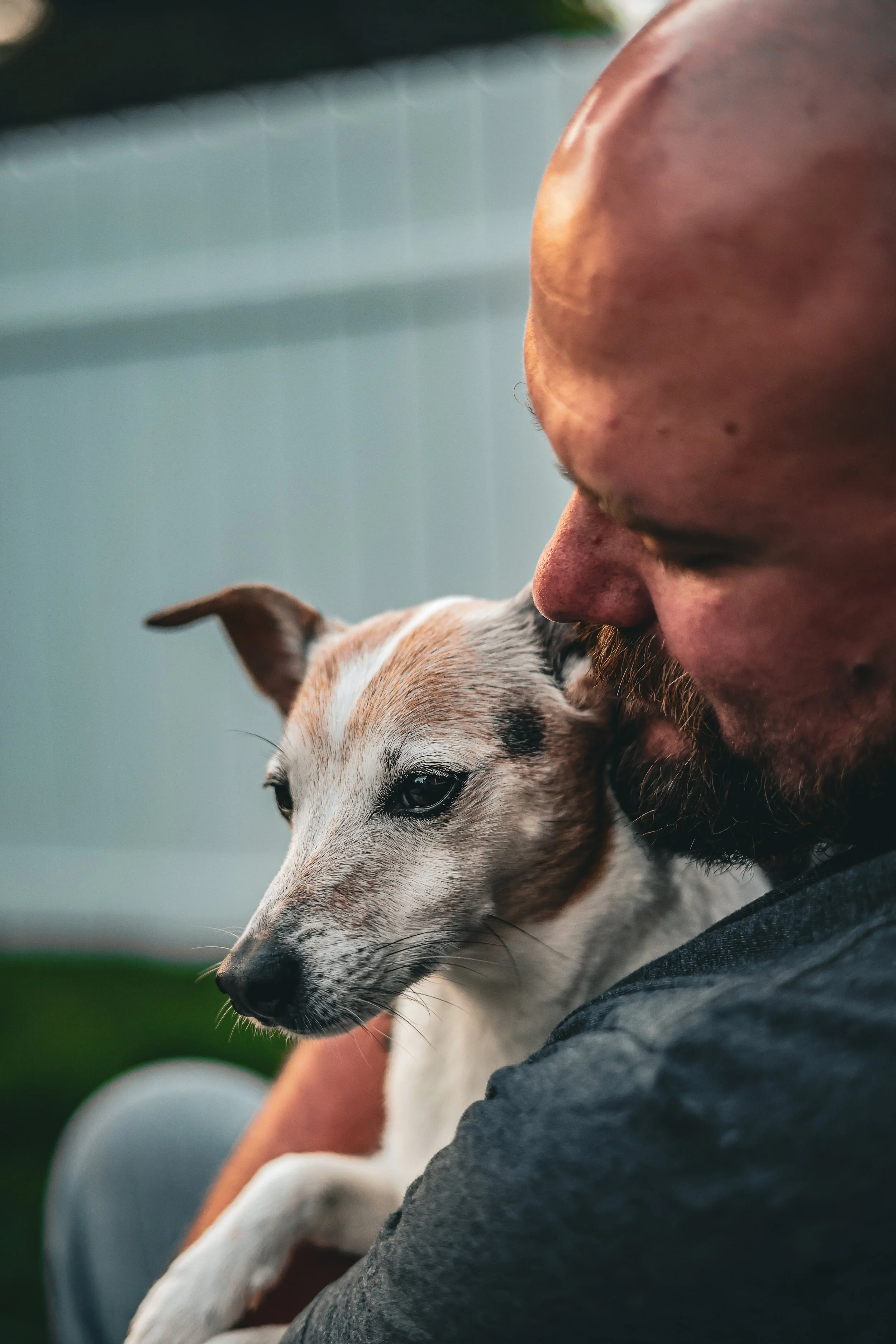A close-up image of a man with a beard and a dog, both with eyes closed, cuddling and resting their heads together, showing a moment of affection and bonding.