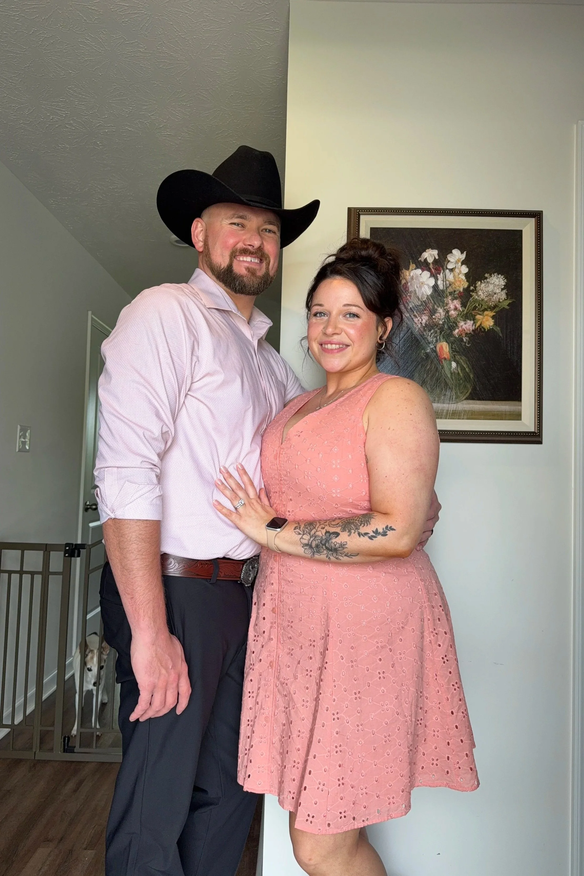 A man wearing a large cowboy hat and a woman with dark hair and a tattoo on her left arm, both smiling and standing close together inside a house in front of a framed floral painting on the wall.