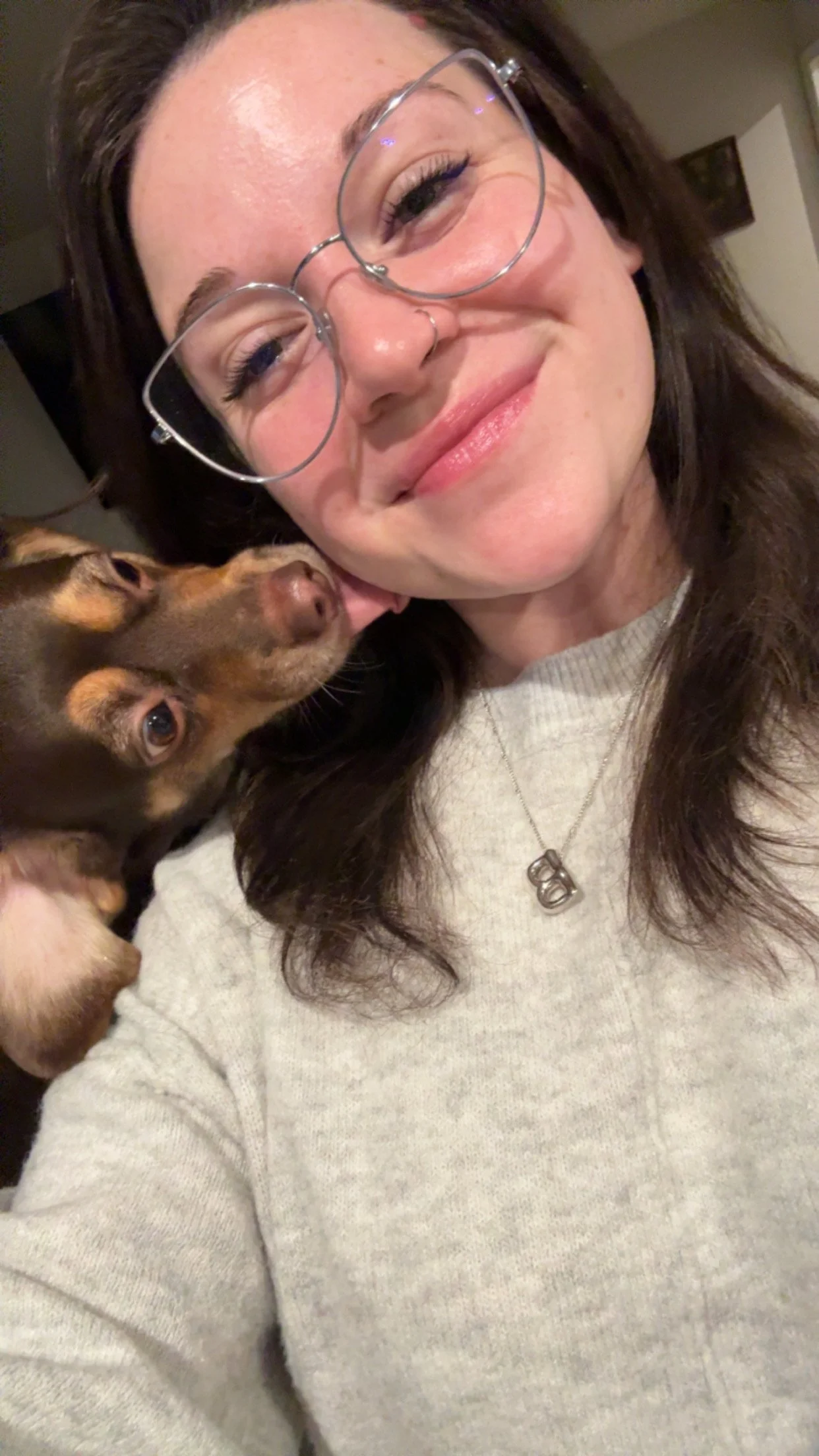A woman with glasses and long brown hair smiling while holding a small brown dog close to her face.