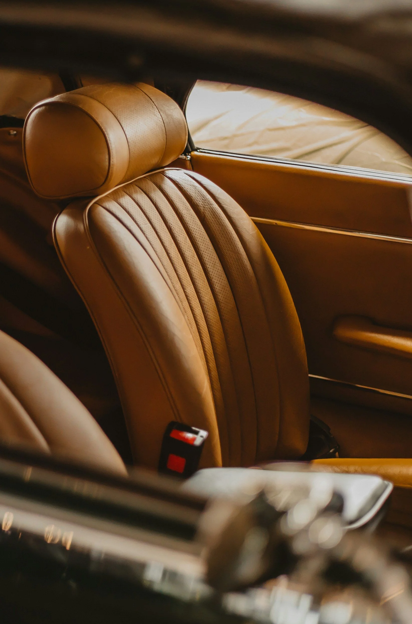 The backseat area of a vintage car with tan leather upholstery, including a headrest and a seatbelt.
