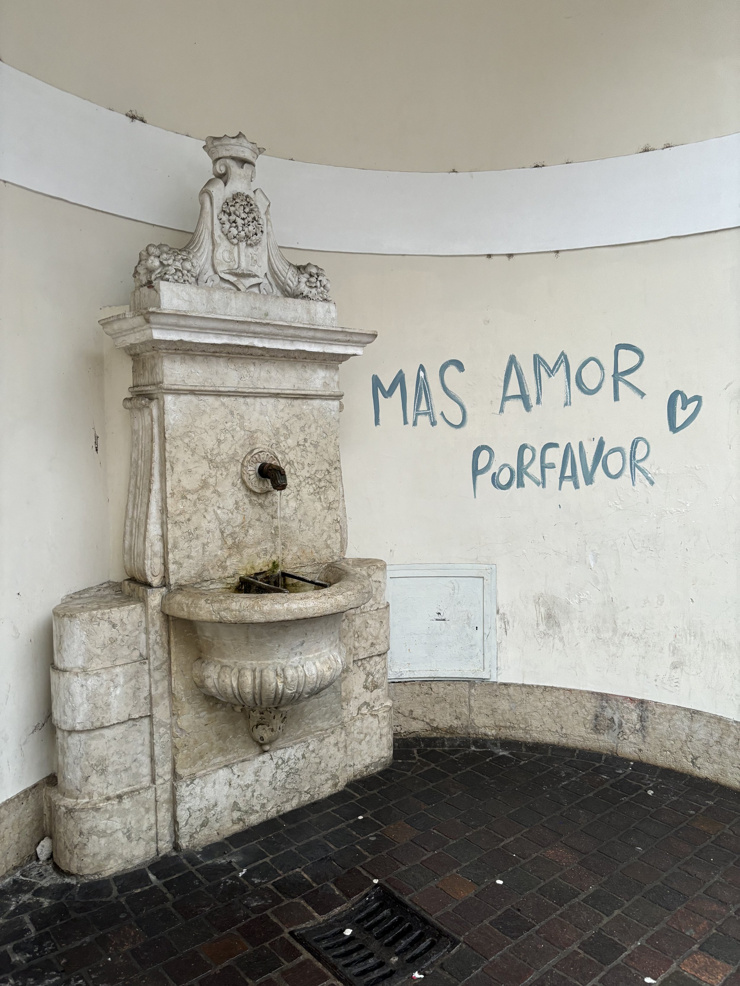 This image depicts a marble fountain in Rovereto, Italy. To the right of it, someone has graffitied "mas amor por favour," which means 'more love please".