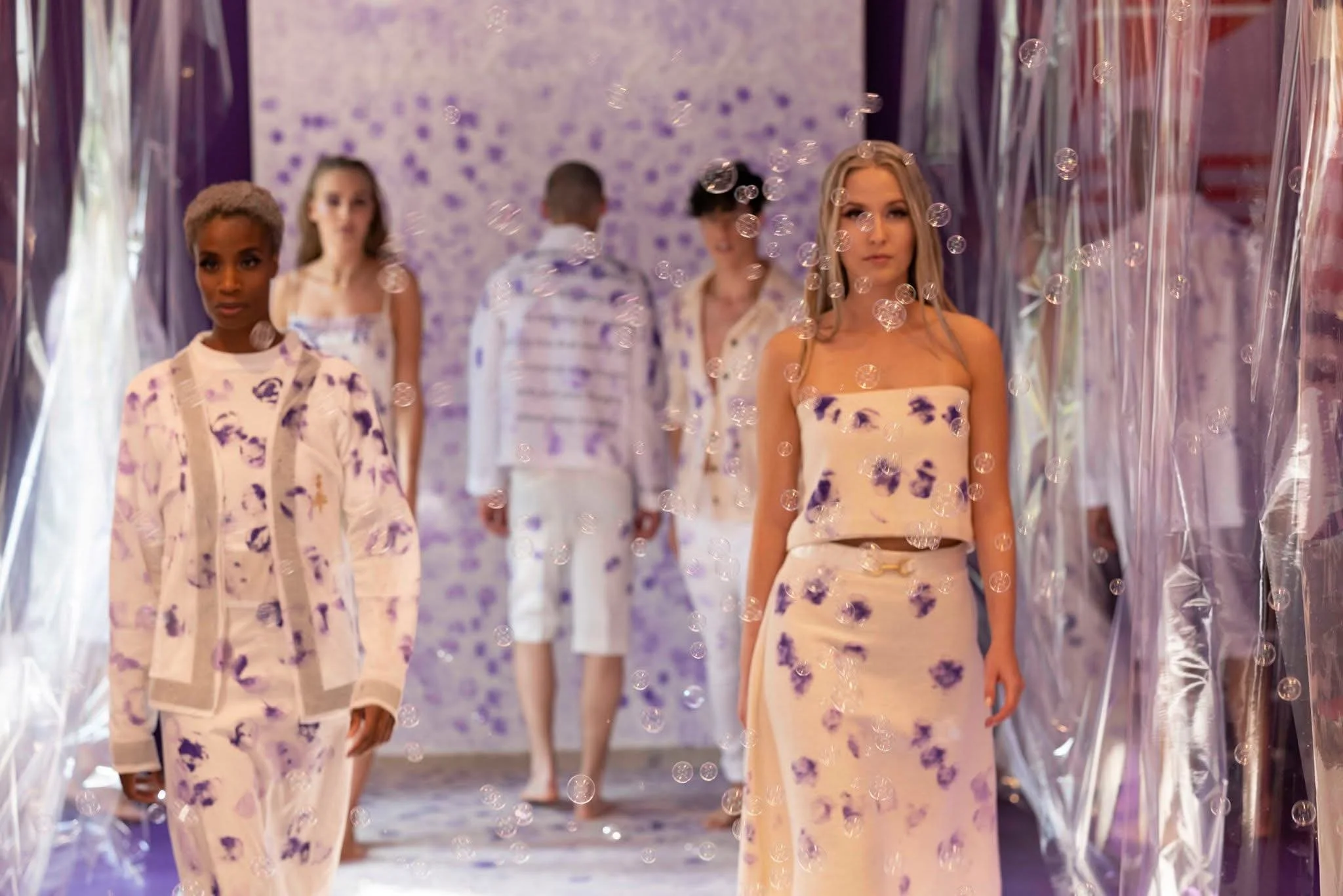 Models walking on a fashion runway wearing matching cream and purple floral outfits, with decorative transparent vertical panels and purple floral decor background.