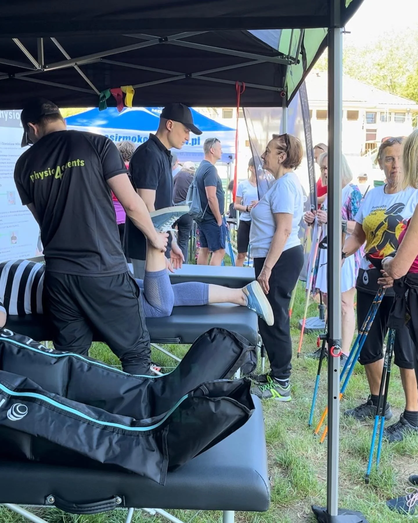 A fitness demonstration at an outdoor event, with a therapist stretching a woman's leg on a massage table under a canopy, surrounded by onlookers and other participants.