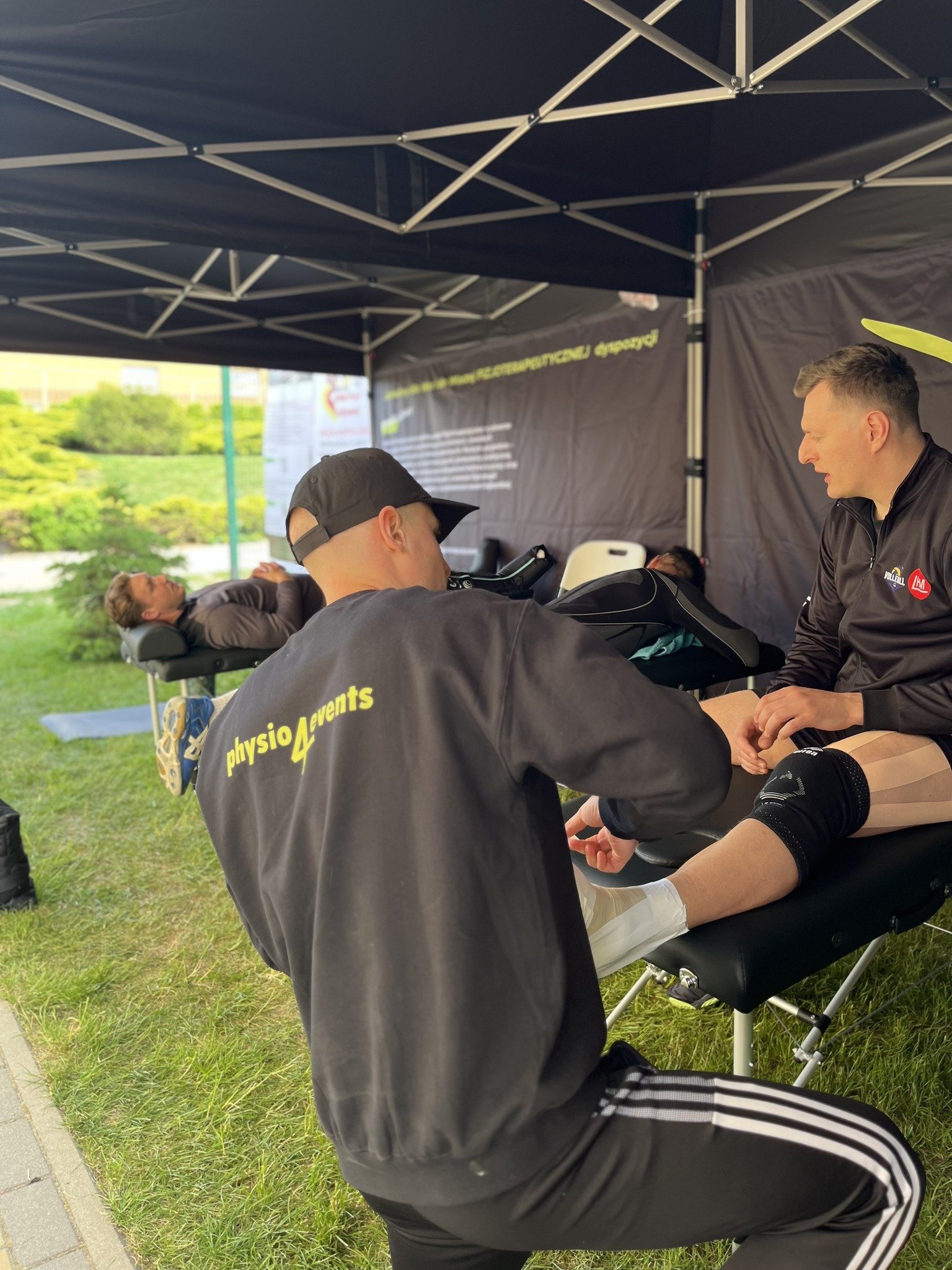 A person receiving a professional leg massage at an outdoor event under a black canopy, with another person lying on a massage table in the background.
