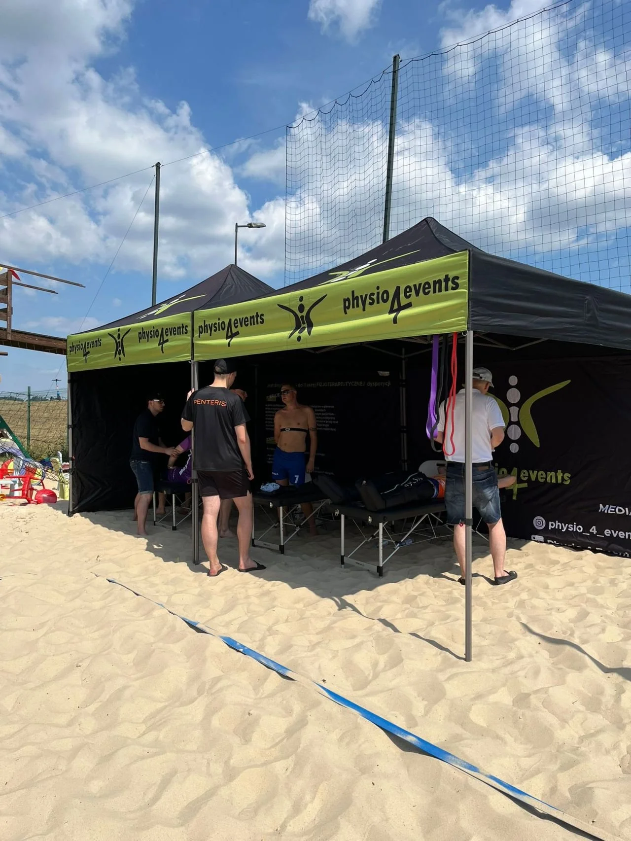 A beach scene with a black tent labeled 'physio4events' and several people inside and around it. The tent is set up on sandy ground with a blue sky and some clouds above.