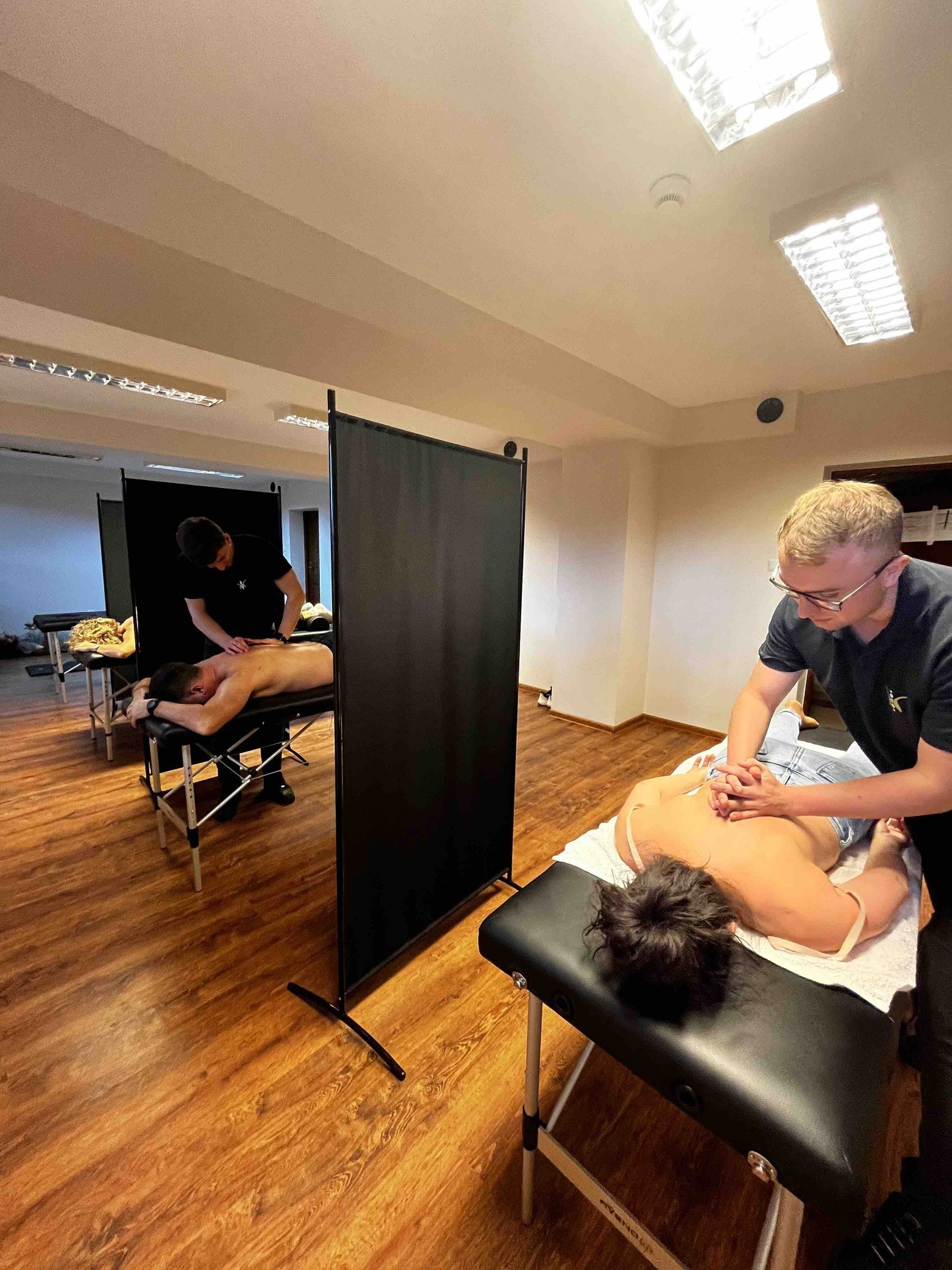 Two massage therapists giving massages to male clients in a massage therapy room divided by black privacy screens. The room has wooden floors and overhead fluorescent lighting.