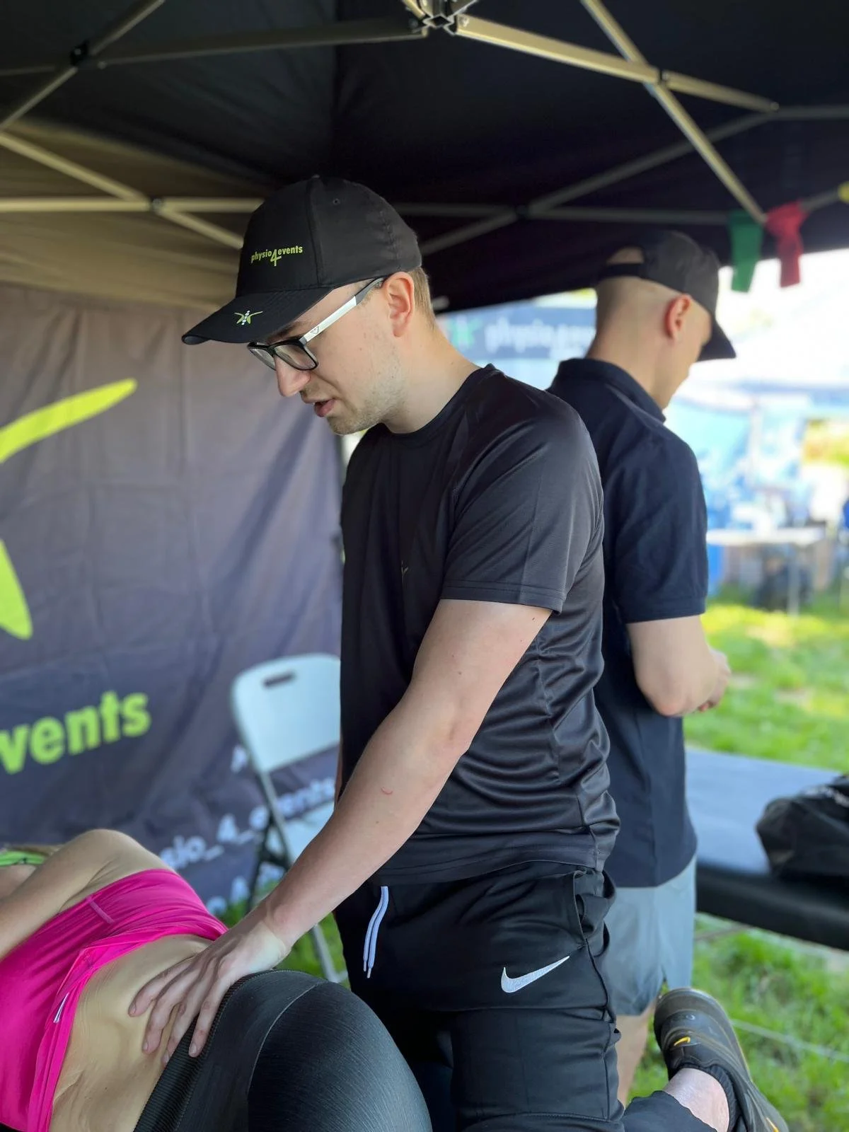 A man wearing glasses, a black cap, and sportswear providing a massage or therapy to a woman lying face down on a massage table at an outdoor event under a canopy.