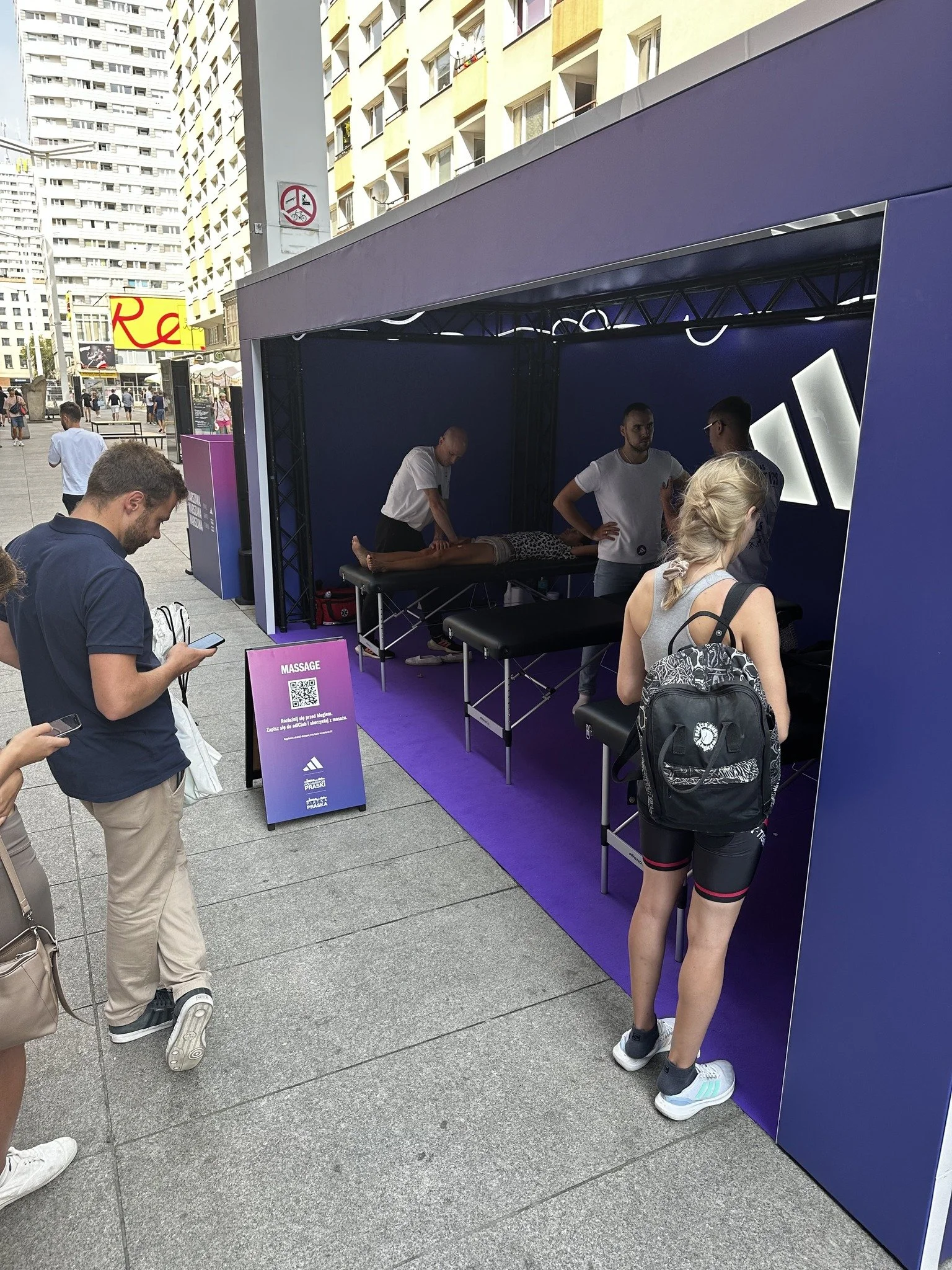 People standing outside a blue promotional tent for massages, with a massage table and staff inside. A sign on a purple stand indicates massage services.