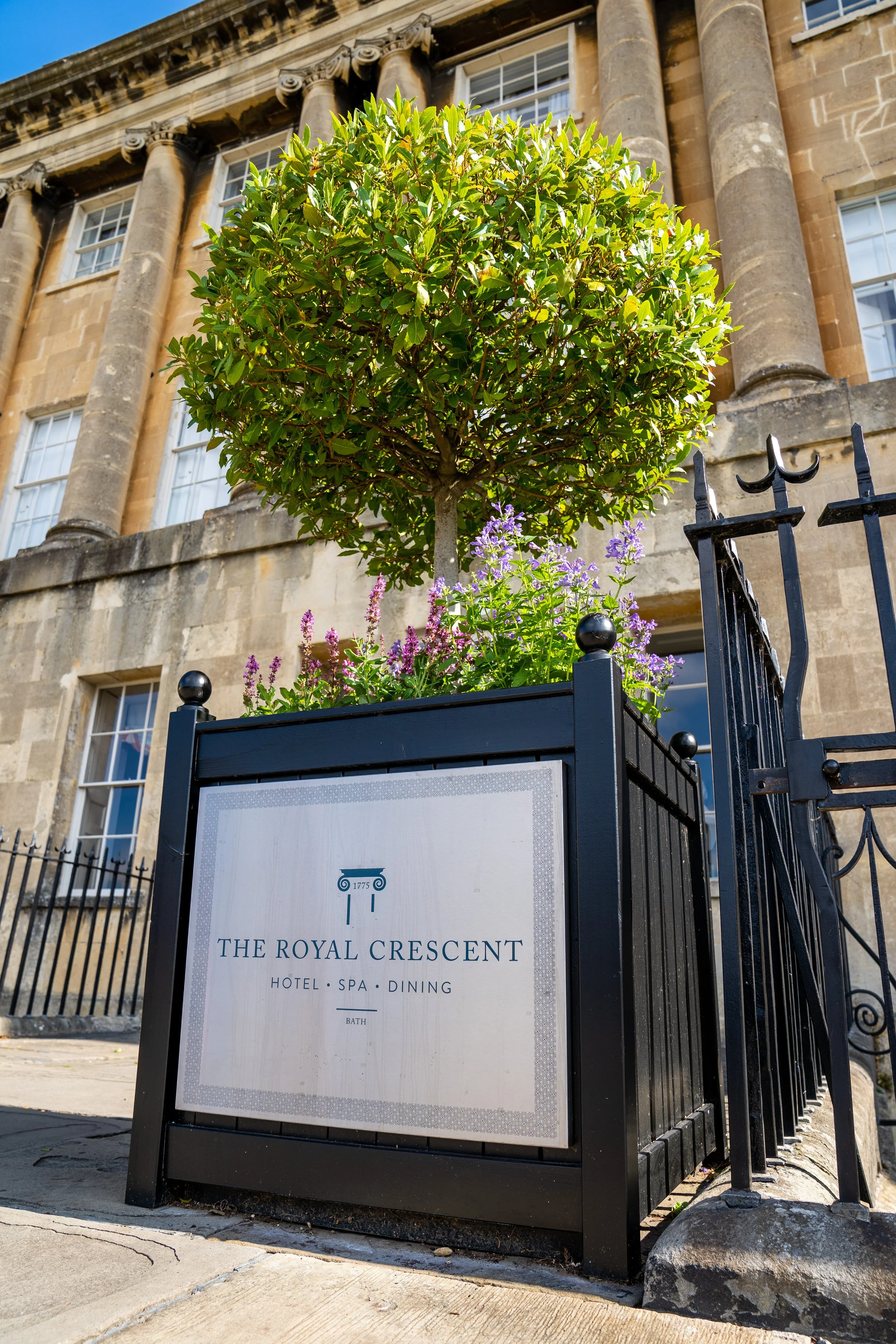 Sign for The Royal Crescent Hotel Spa Dining with logo, black frame, and greenery in front of a historical building with large windows and stone columns.
