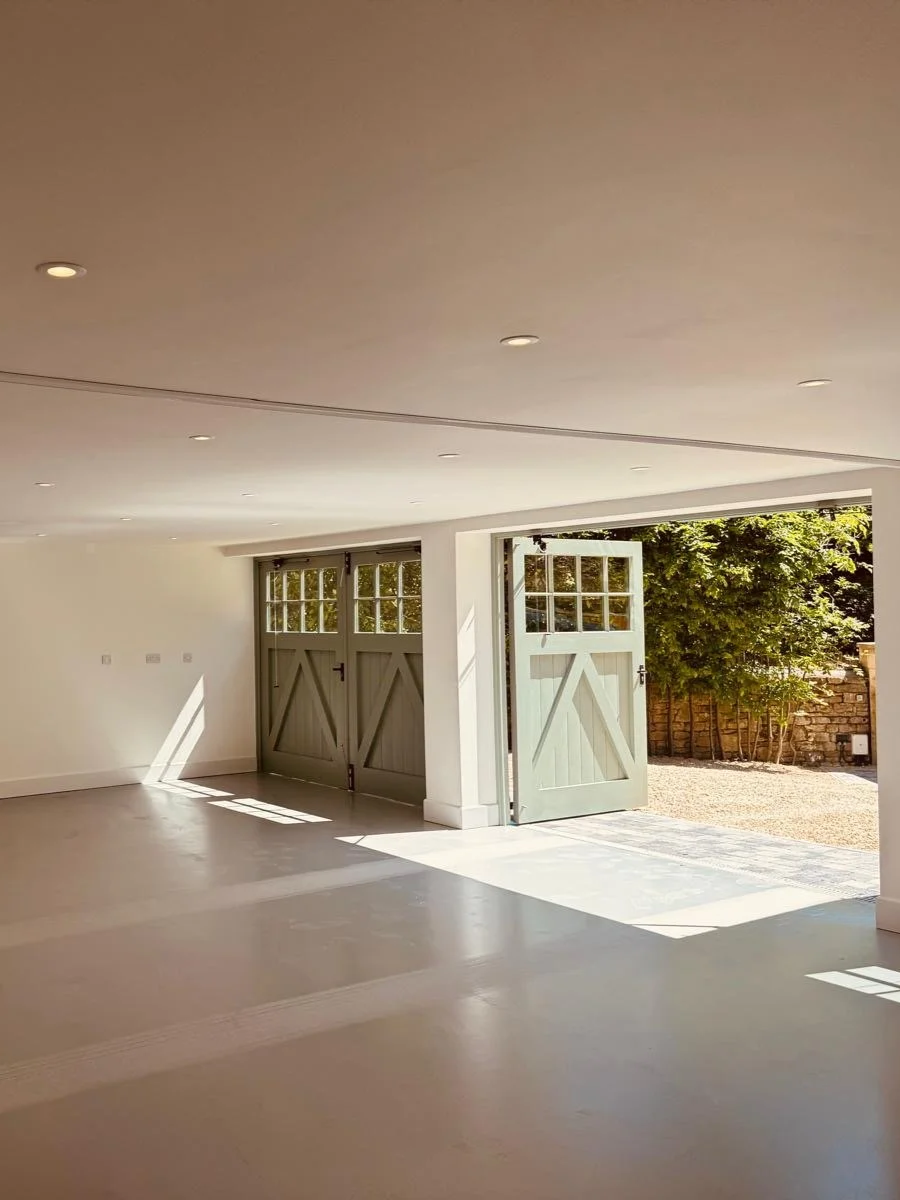 Empty interior room with open double doors leading outside to a garden yard, sunlight casting shadows on the floor.
