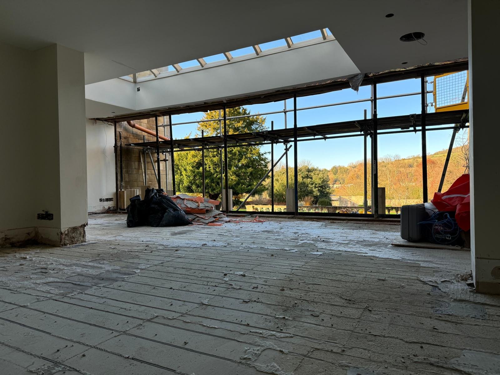 Interior of a room under construction or renovation, with large window openings, construction materials, and debris on the floor, and scaffolding outside.