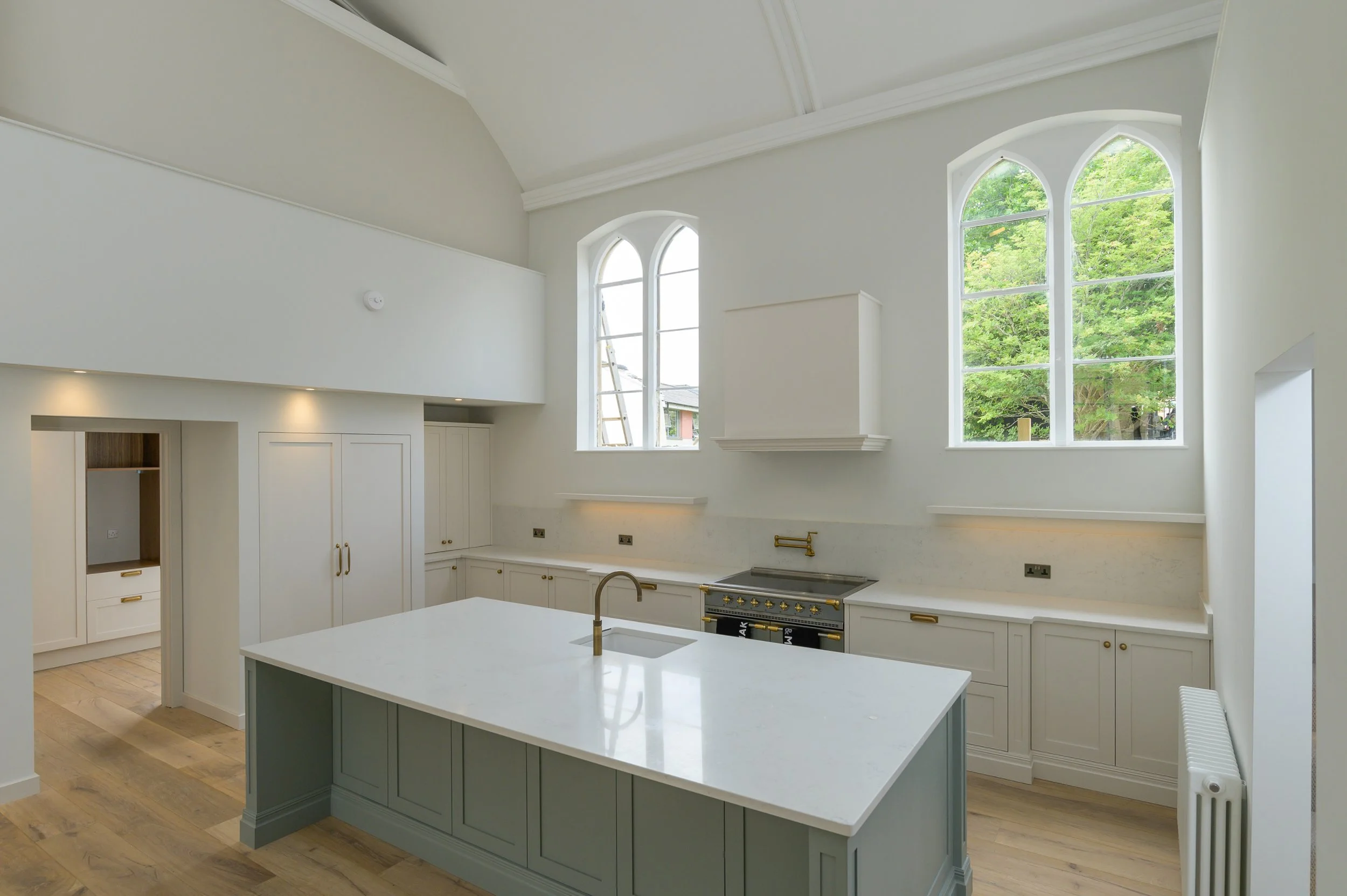 Modern kitchen with white cabinetry, a large kitchen island with grey base, and high arched windows showing green trees outside.