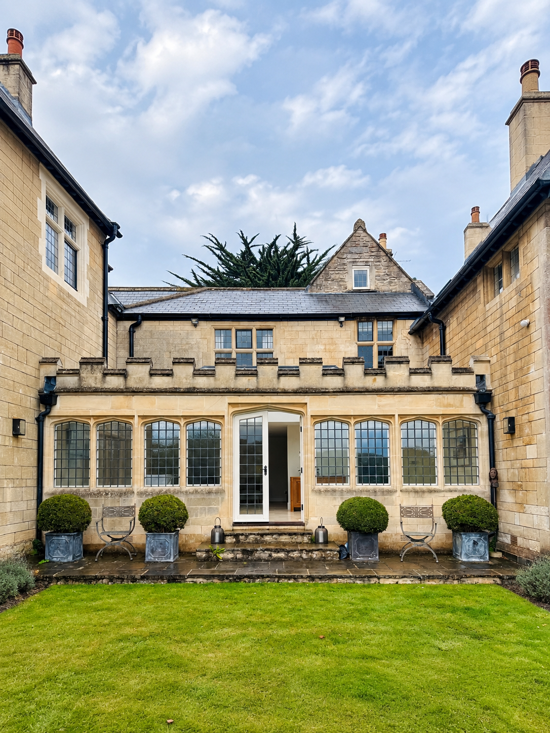 Photo of a large house with stone walls and multiple windows, a glass sunroom at the back, and a well-maintained grassy lawn under a bright blue sky.