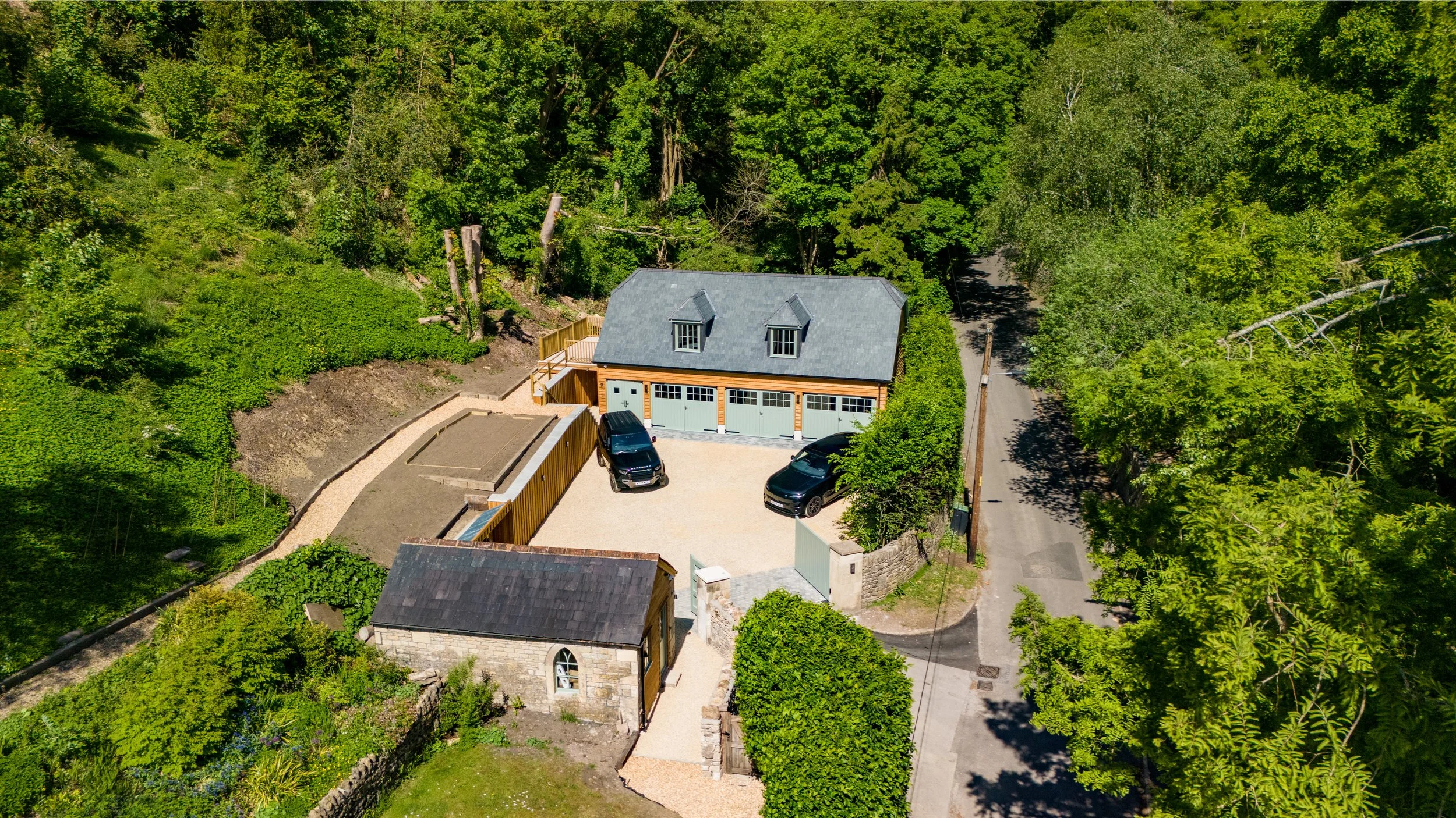 A two-story house with a gray roof and large garage doors, surrounded by green trees, with a driveway and two black cars parked in front.