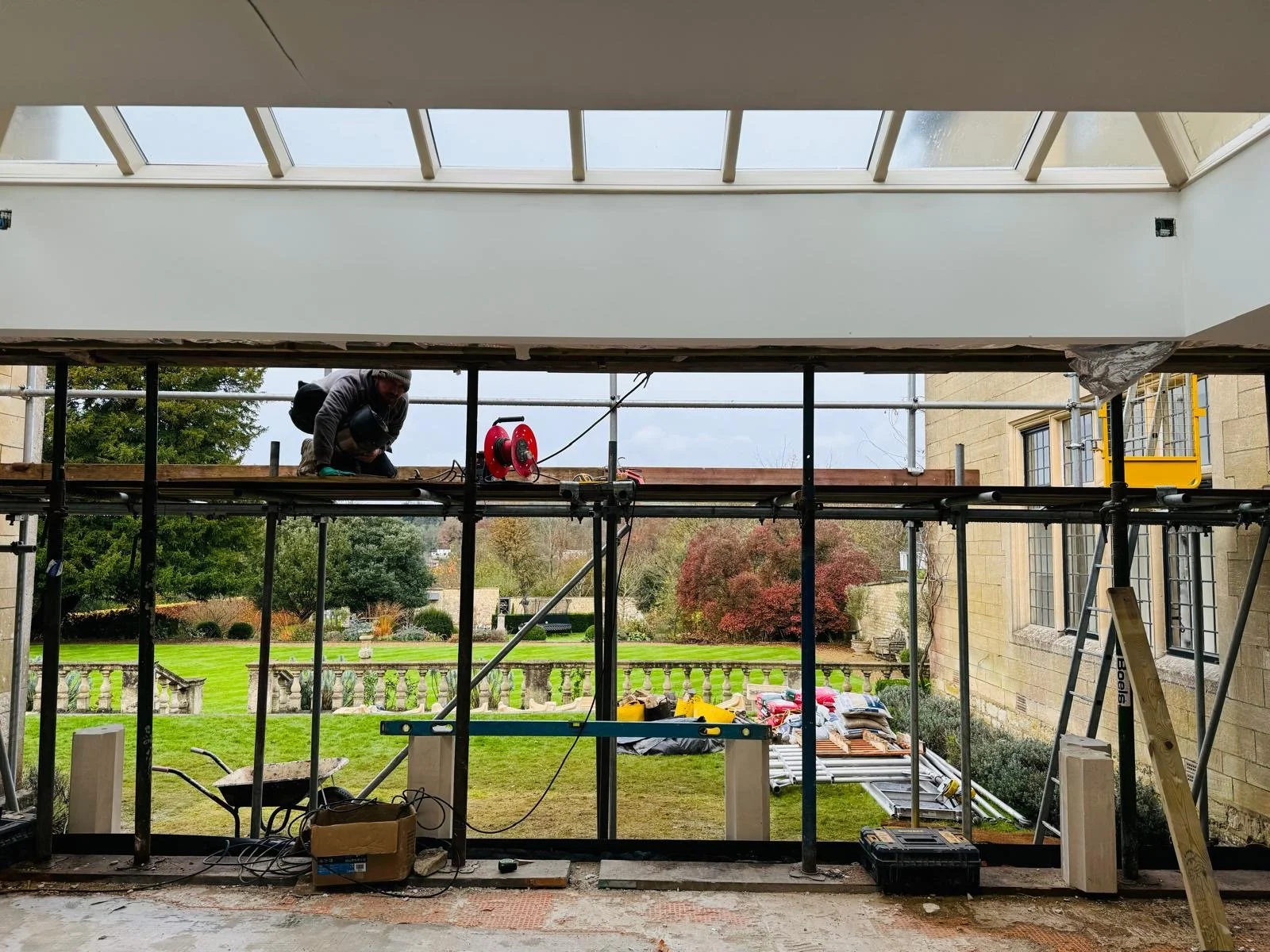Construction worker working on the upper level of a building under construction, with a view of the backyard and trees outside.