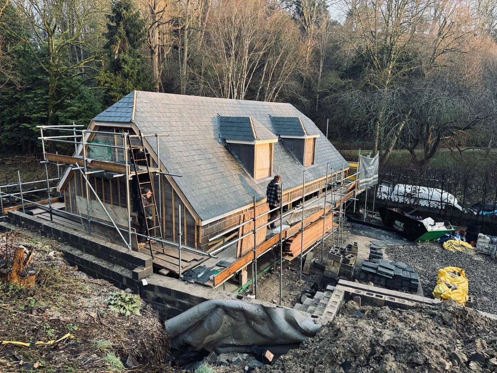 Construction workers building a house with a steep roof, scaffolding around the structure, and a wooded background.