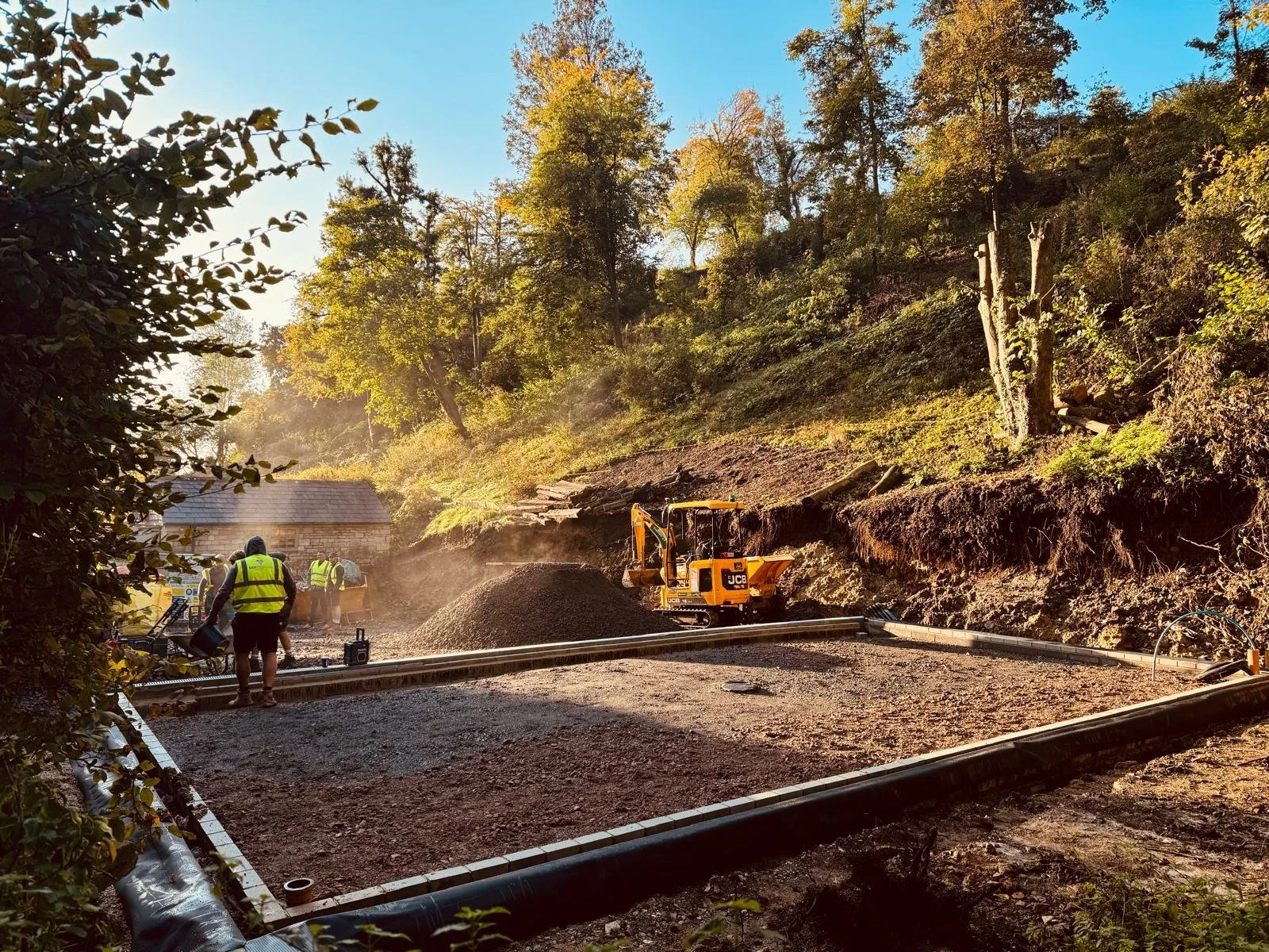 Construction workers in high-visibility vests working on a road repair in a wooded area, with an excavator and construction materials visible.