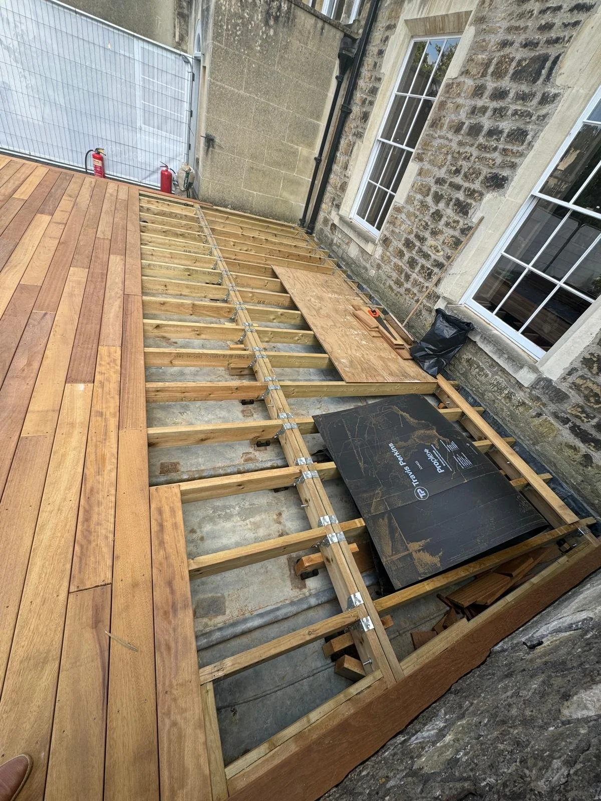 Partial wooden deck construction with some planks already installed and other sections showing the underlying framework of wooden beams and supports, next to a stone building with white-framed windows.