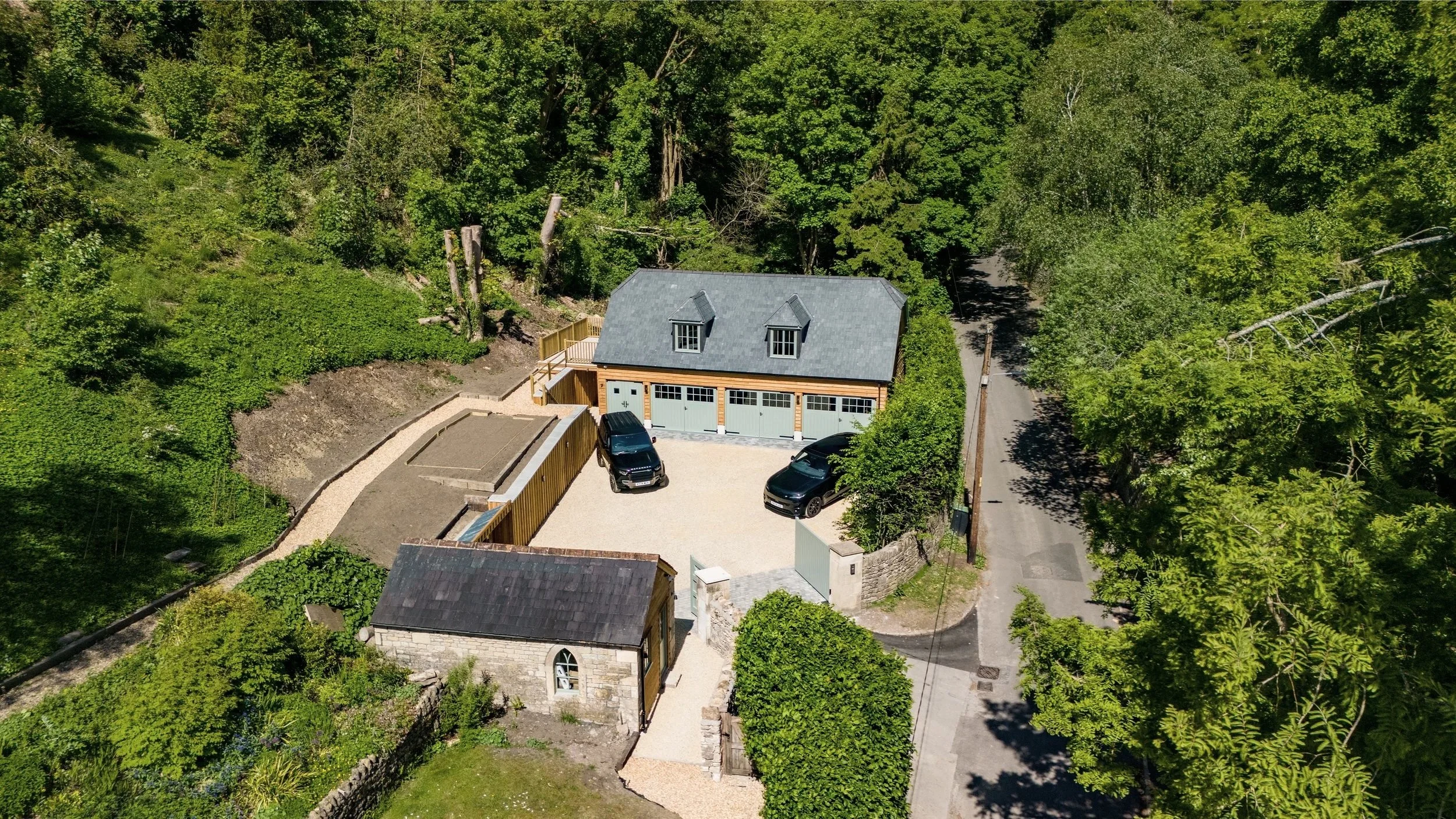 Aerial view of a residential property with a modern newly built garage, two black cars, surrounded by lush green trees.