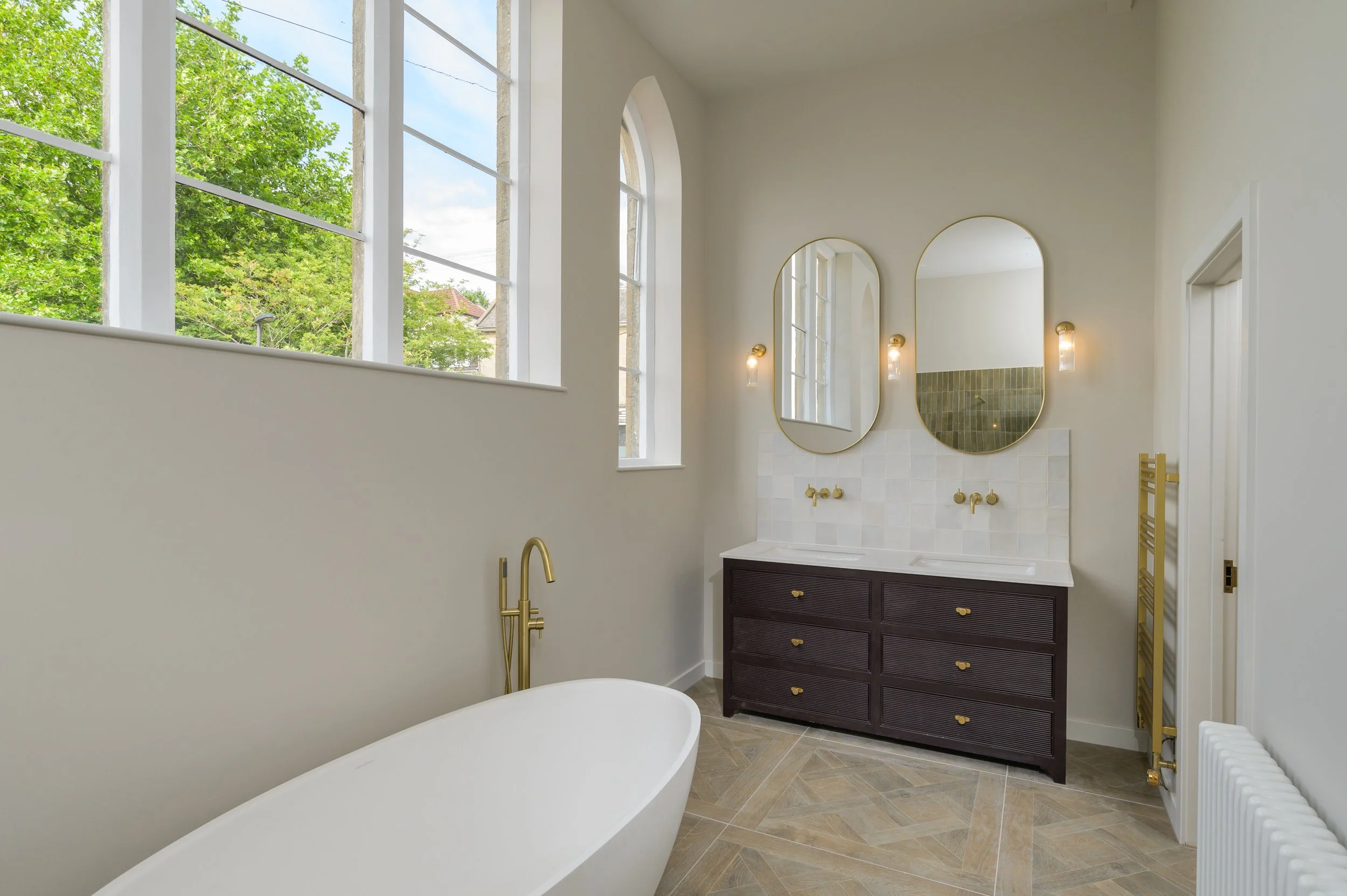 Bright bathroom with large windows showing green trees outside, featuring a white bathtub, gold fixtures, dark wood vanity with gold handles, two mirrors with gold frames, and yellow towel racks.