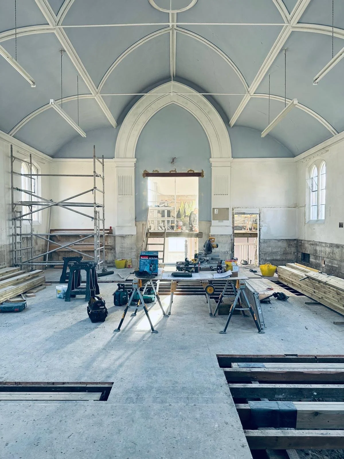 Interior of a building under renovation, with construction tools, scaffolding, stacks of wood, and a partially removed floor revealing wooden beams.