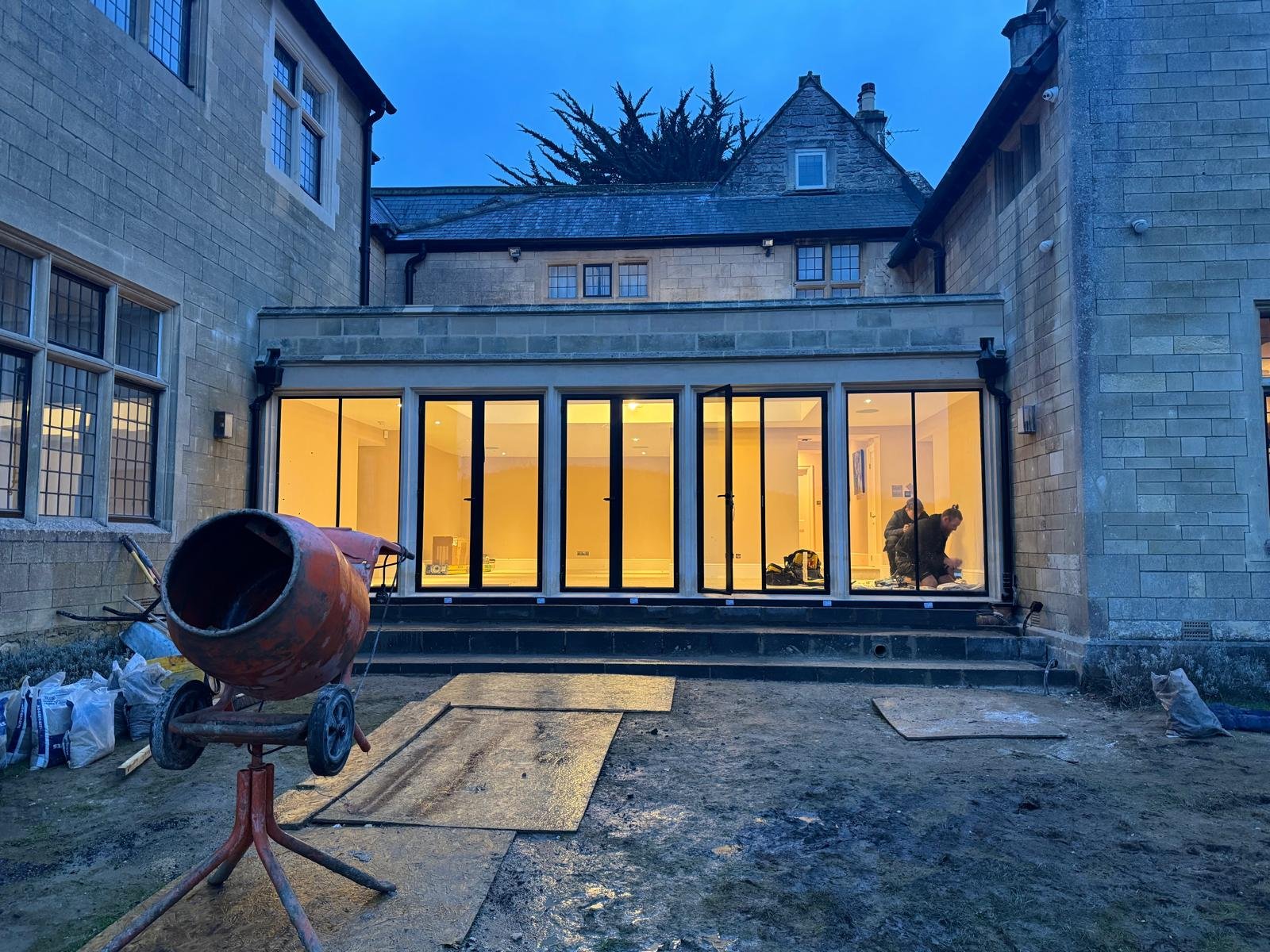 Construction site of a new glass-enclosed extension on a brick house at dusk, with two workers inside working, and construction equipment and materials outside.