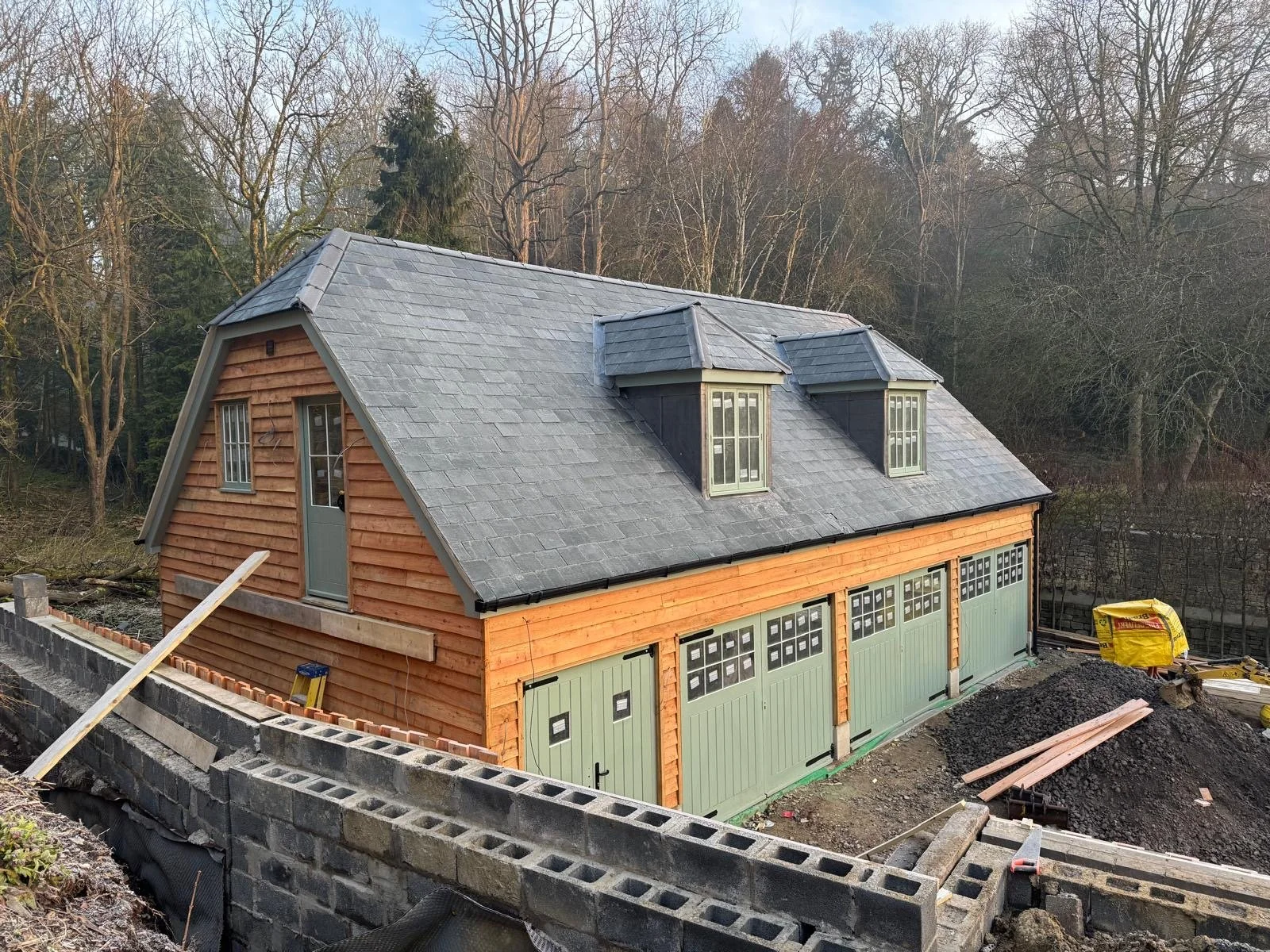A wooden house under construction with a sloped roof and multiple windows, surrounded by trees and construction materials.