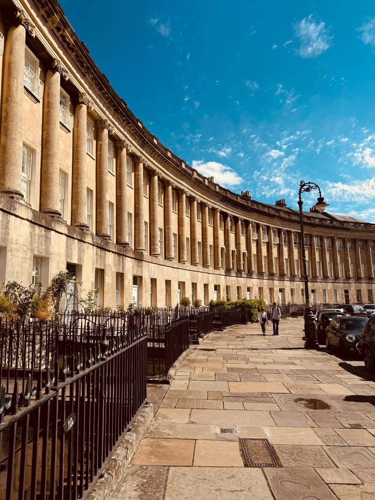 Curved historic building with classical columns, a street with parked cars, a black iron fence, a street lamp, and two people walking on the sidewalk under a blue sky with some clouds.