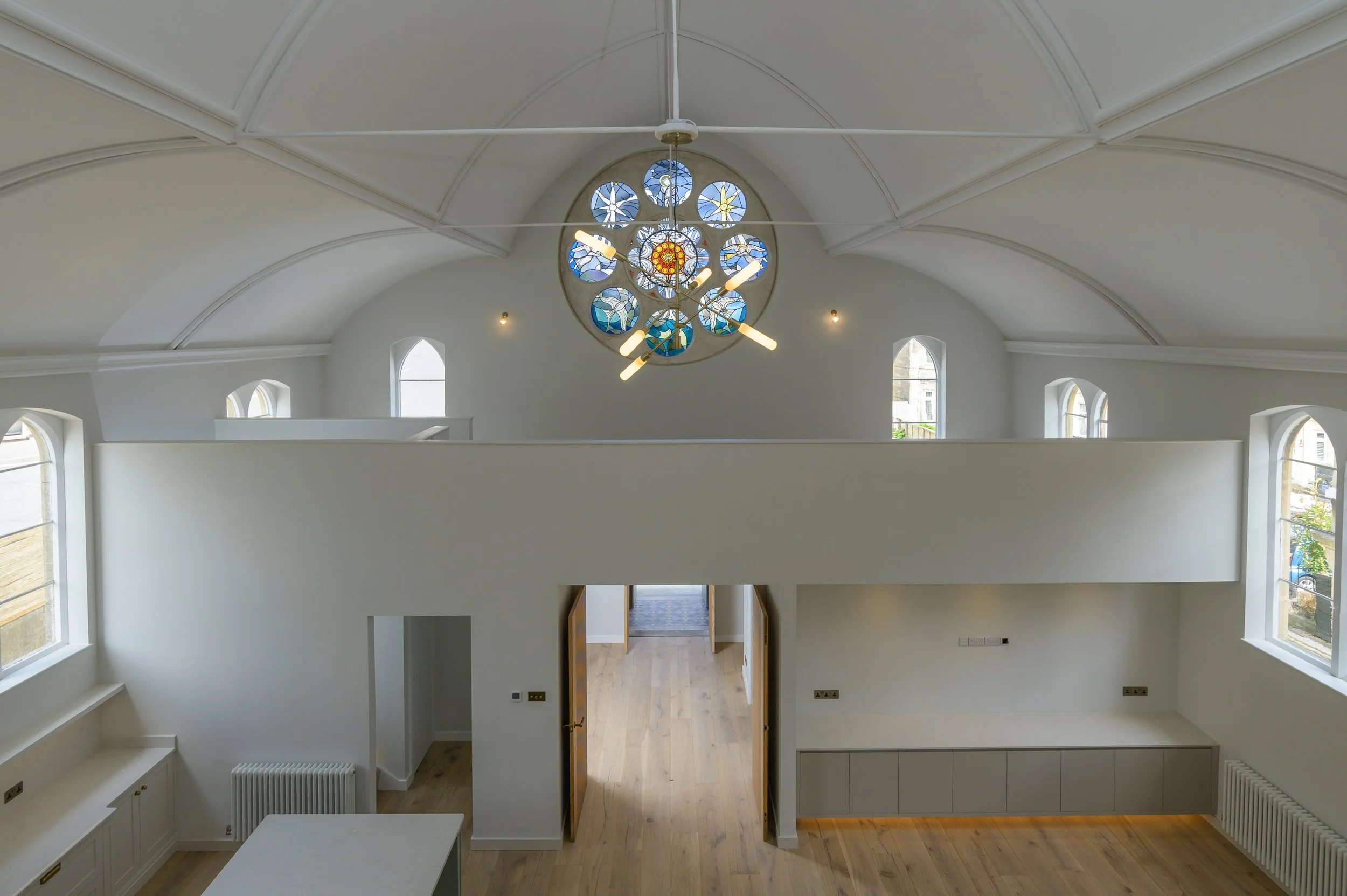 Interior of a modern, white-walled building with a high, vaulted ceiling. There is a large circular stained glass window with blue and yellow patterns, and a contemporary chandelier hanging in front of it. Multiple arched windows let in natural light, and the space includes a mezzanine or upper level with a white railing.