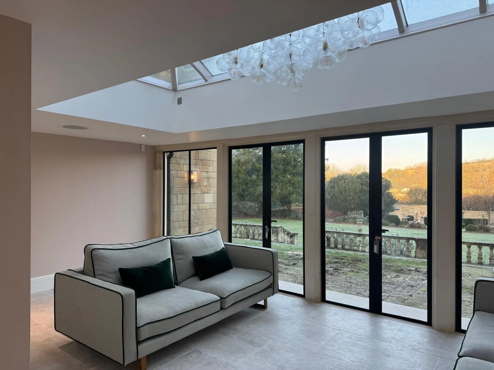Modern living room with large floor-to-ceiling glass doors and a skylight, overlooking a garden and trees.