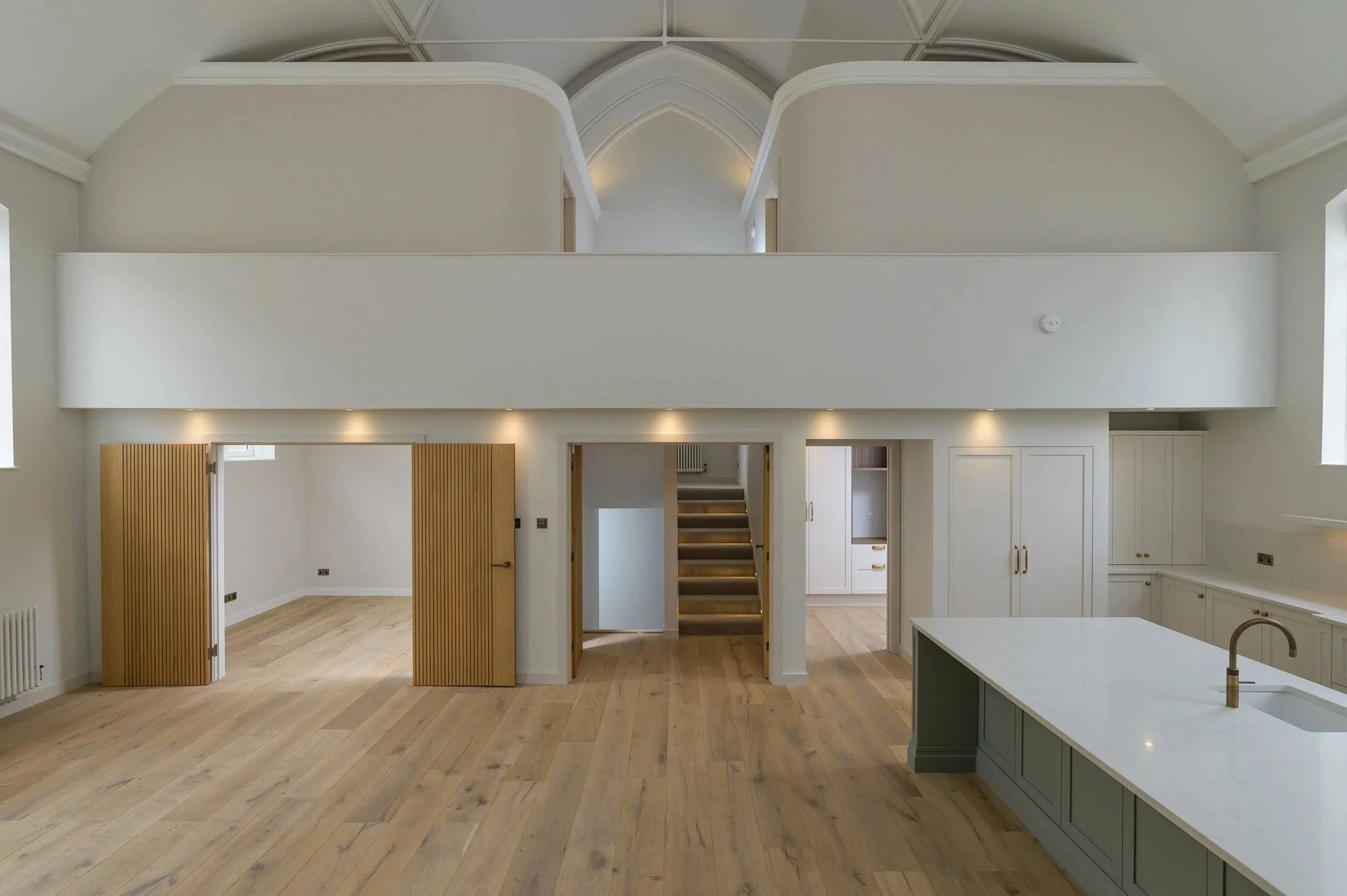 Interior view of a modern, spacious kitchen with hardwood floors, white cabinetry, and a kitchen island with a gray base. There are wooden doors that slide open to reveal additional rooms, and a staircase leading to an upper level with a ceiling featuring arched design elements.