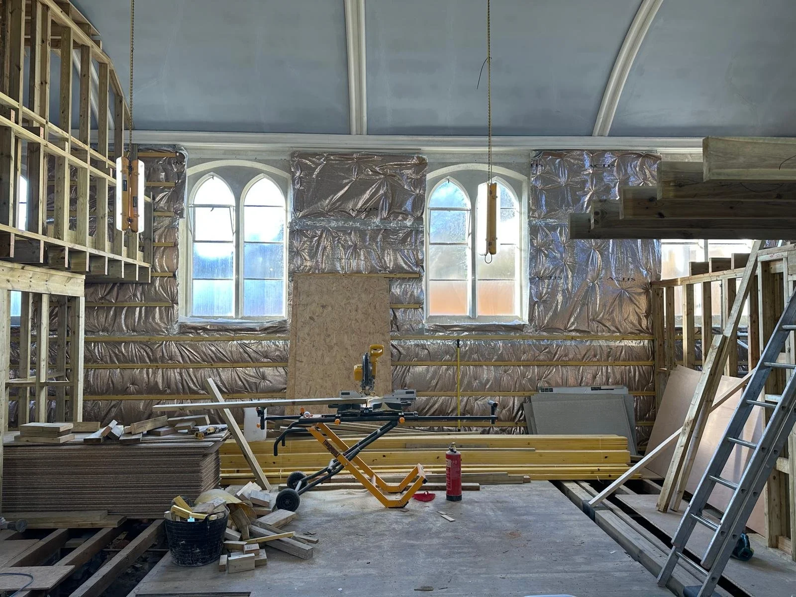 Interior of a building under construction with wooden framing, insulation material, construction tools, and ladders, with large arched windows in the background.